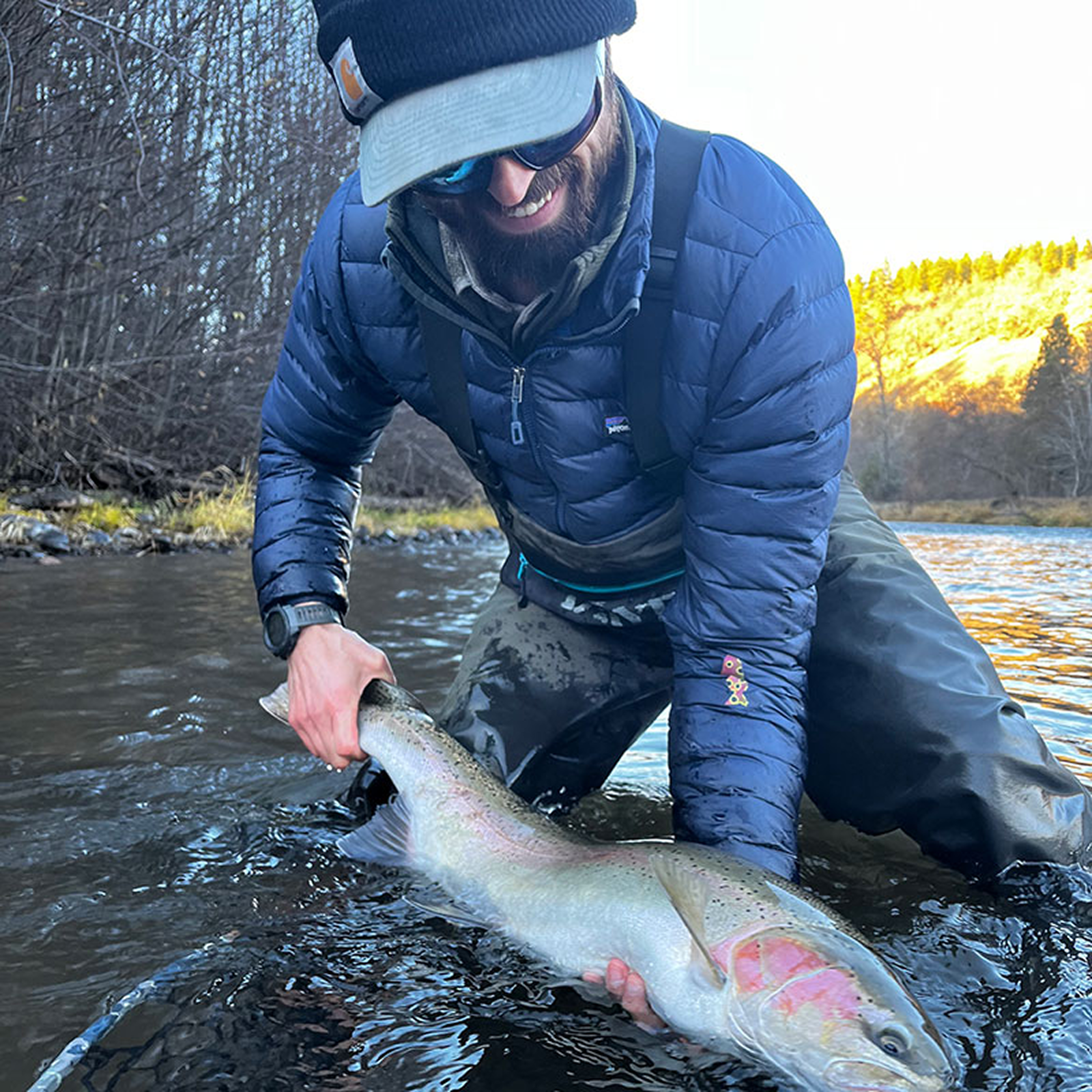 A man wearing a blue jacket, black sunglasses, and a black cap is smiling while holding a large fish in a river during sunset.