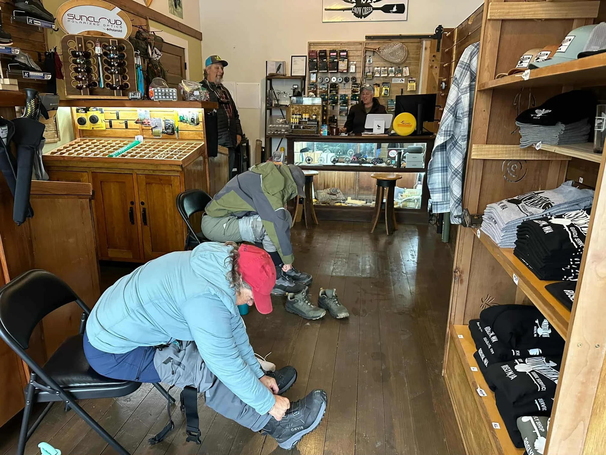 Inside Crow Creek Fly Shop, two people are seated on black chairs near the front, adjusting fishing boots. The store sells fly fishing gear, with hats, shirts, and accessories displayed on shelves and racks.
