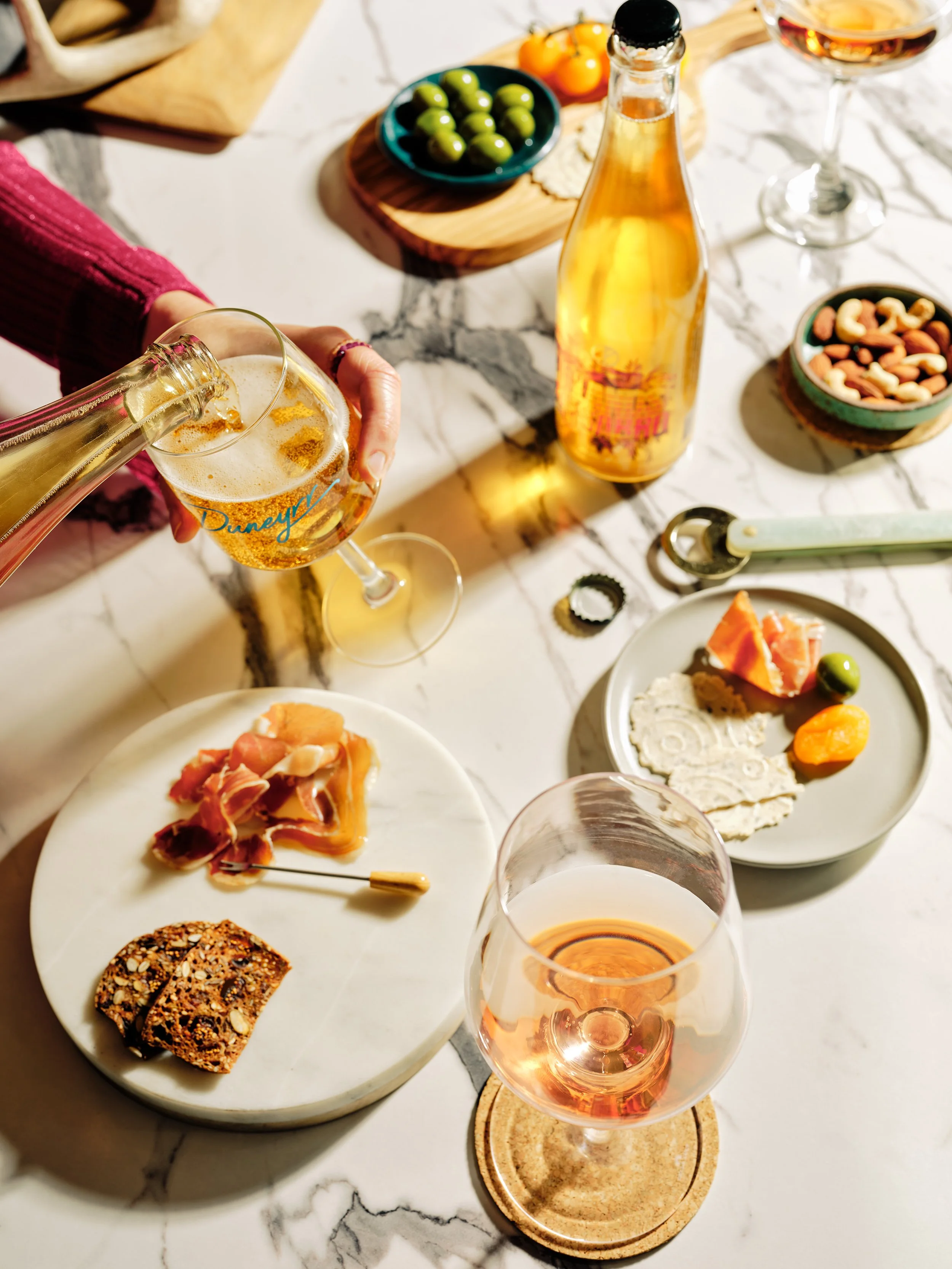 A charcuterie board with various cheeses, cured meats, a cherry tomato, and crackers, a glass of white drink with ice and fruit slices, and a small bowl of fig jam on a marble table.