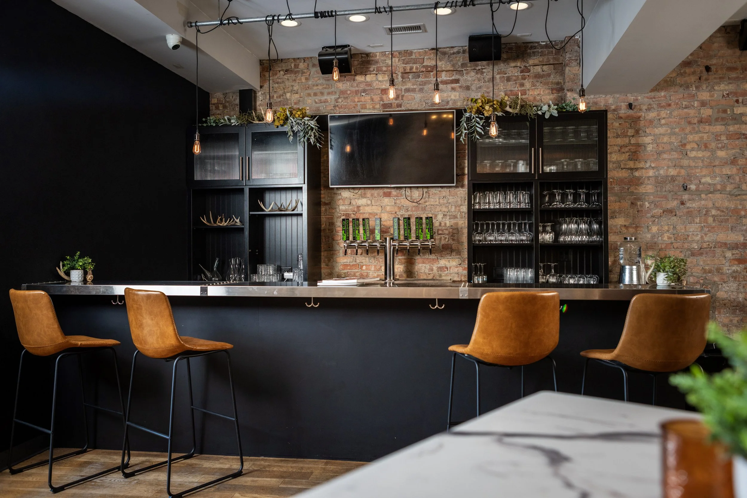 Empty bar area with brown leather chairs, a black and stainless steel counter, brick wall, hanging pendant lights, and additional seating in the background.