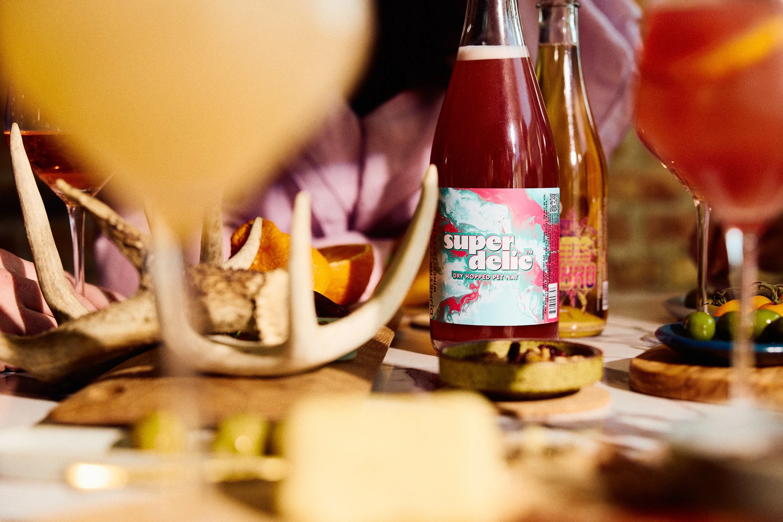 Colorful bottles, food, and decorative items on a table, including a pink bottle labeled 'super deka' and antlers, with blurry glasses and platters of food at Tethered Events