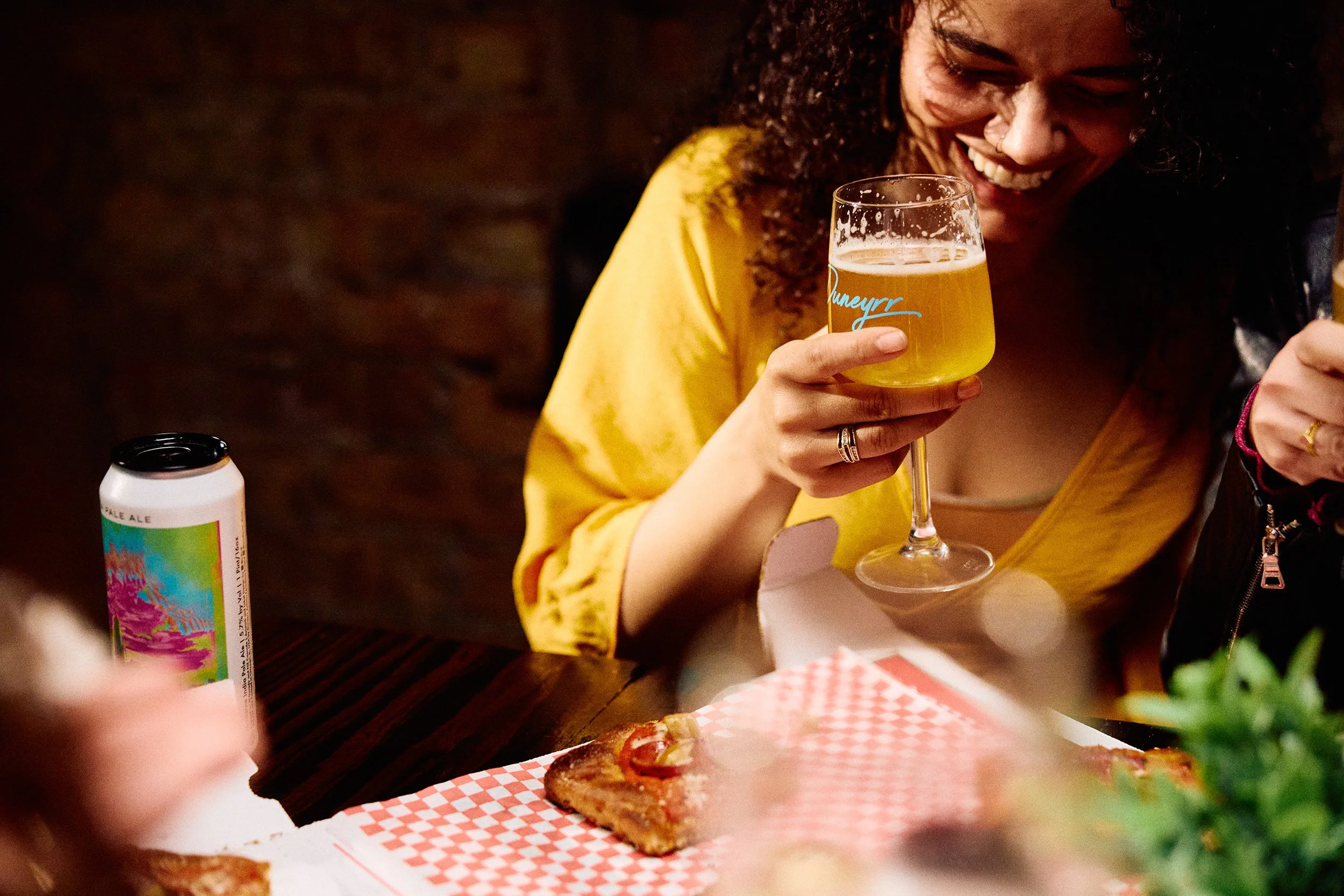 A woman with curly hair wearing a yellow shirt and a nose ring, smiling and holding a glass of beer, sitting at a table with pizza and a can of beer, in a cozy indoor setting.