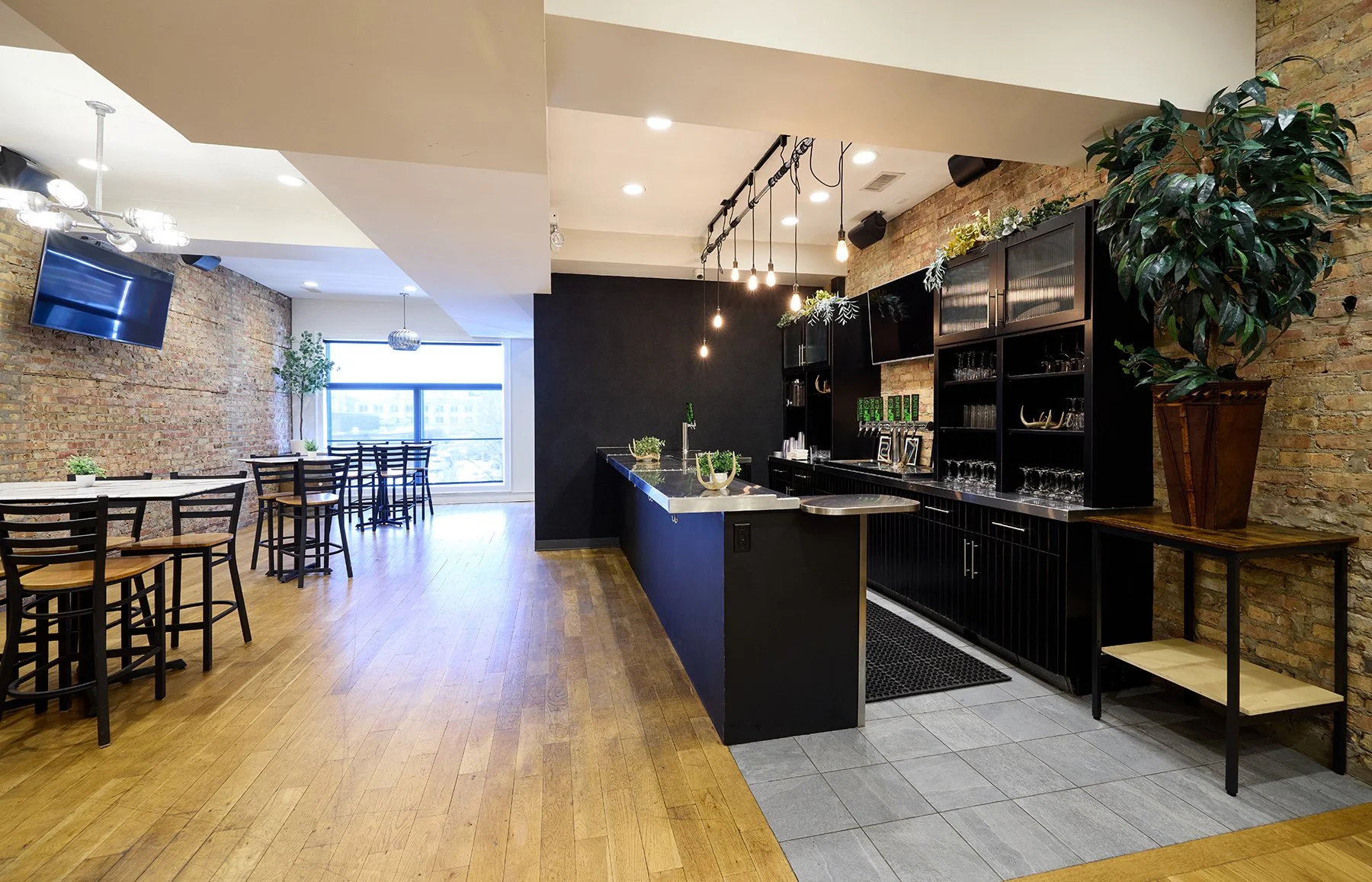 Interior of a modern bar in an event space with a black counter, exposed brick walls, wooden and tile flooring, and seating area with tables and chairs near large window.