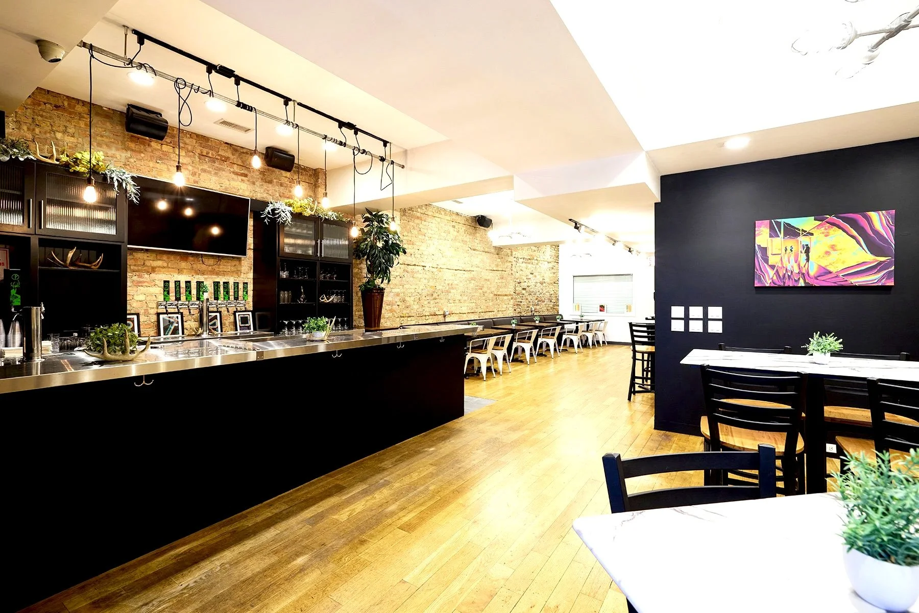 Interior of a modern event space with a long bar counter, brick walls, hanging light bulbs, and black and white chairs around tables decorated with small plants.
