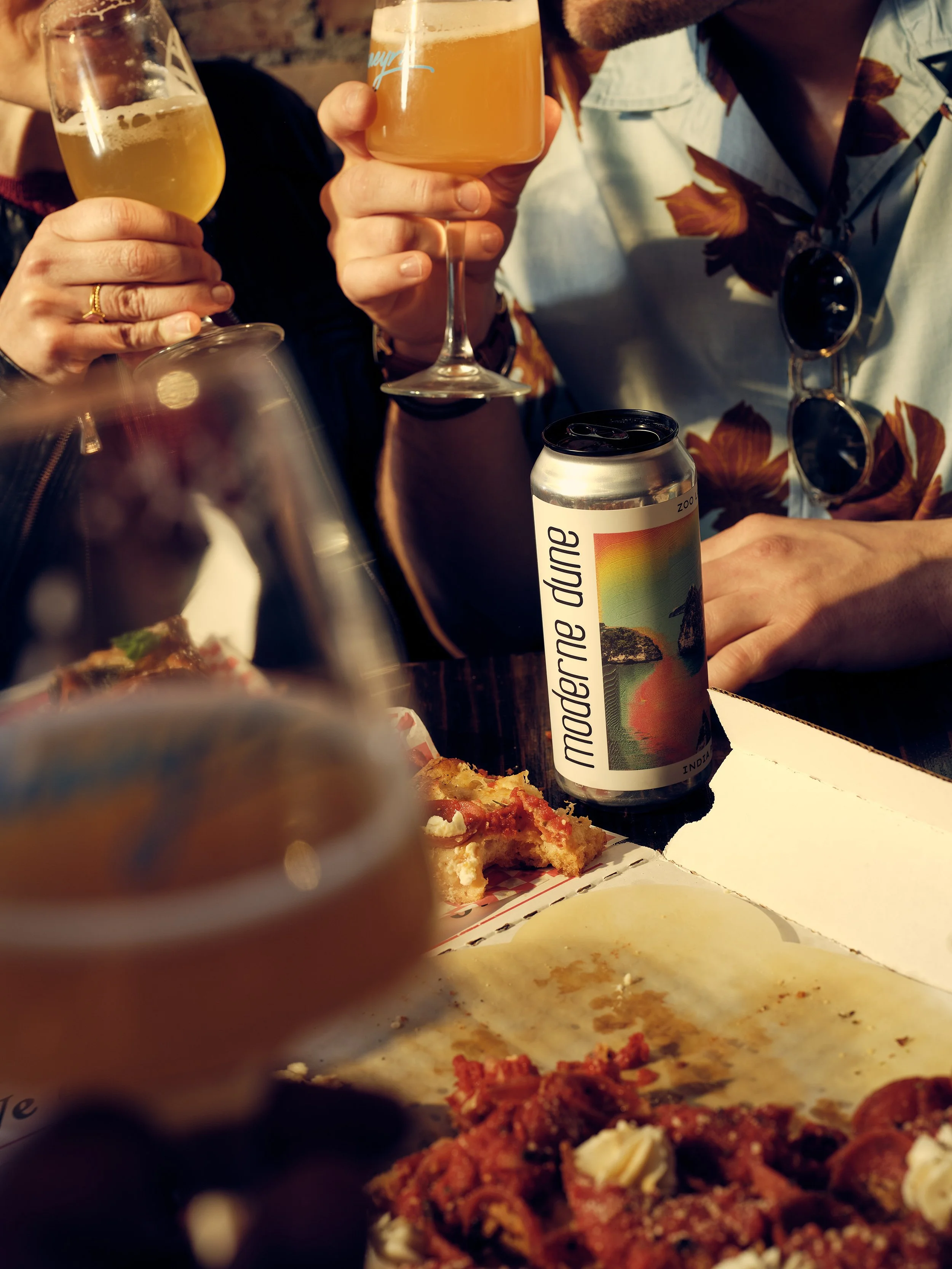 People clinking glasses of beer during a casual gathering, with food and a modern dune beverage can on the table.