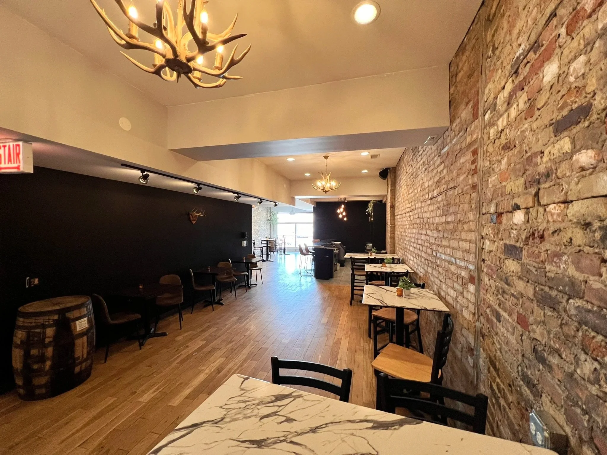 Interior view of a modern dining and lounge area with wood flooring, a brick wall, black benches and chairs, small potted plant on table, colorful artwork on black wall, antler chandelier, hanging pendant lights, and large windows at the far end.