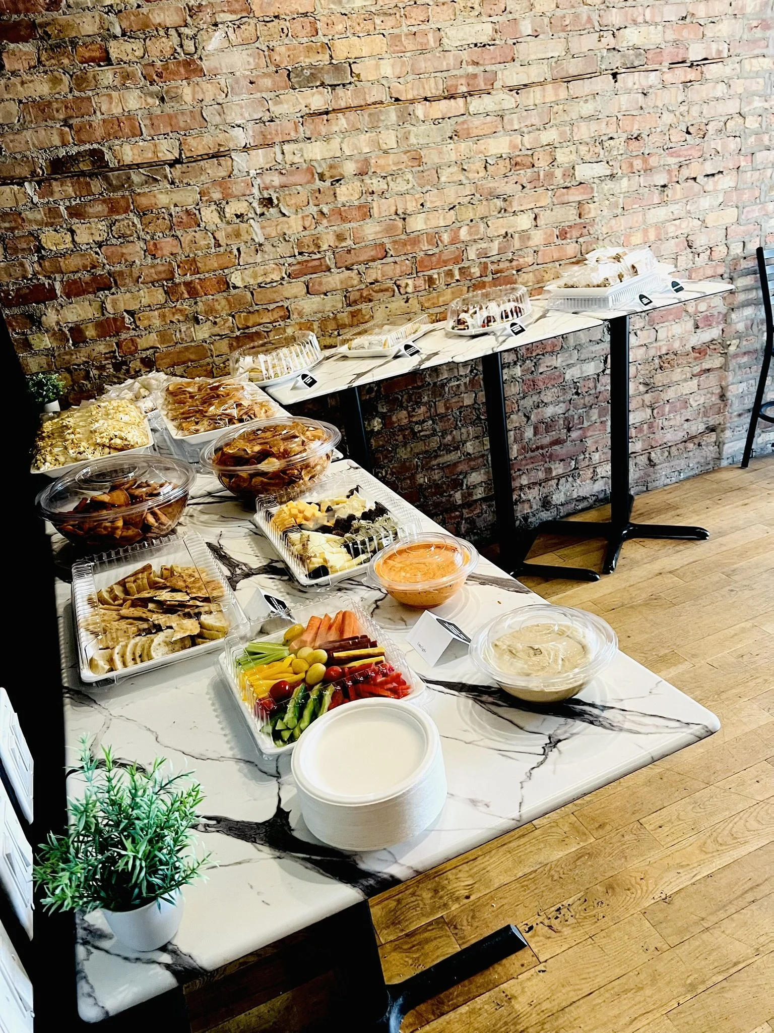 Buffet table with various dishes, including cheese, fruit, vegetables, dips, and garnishes, set against a brick wall with some trays and plates.