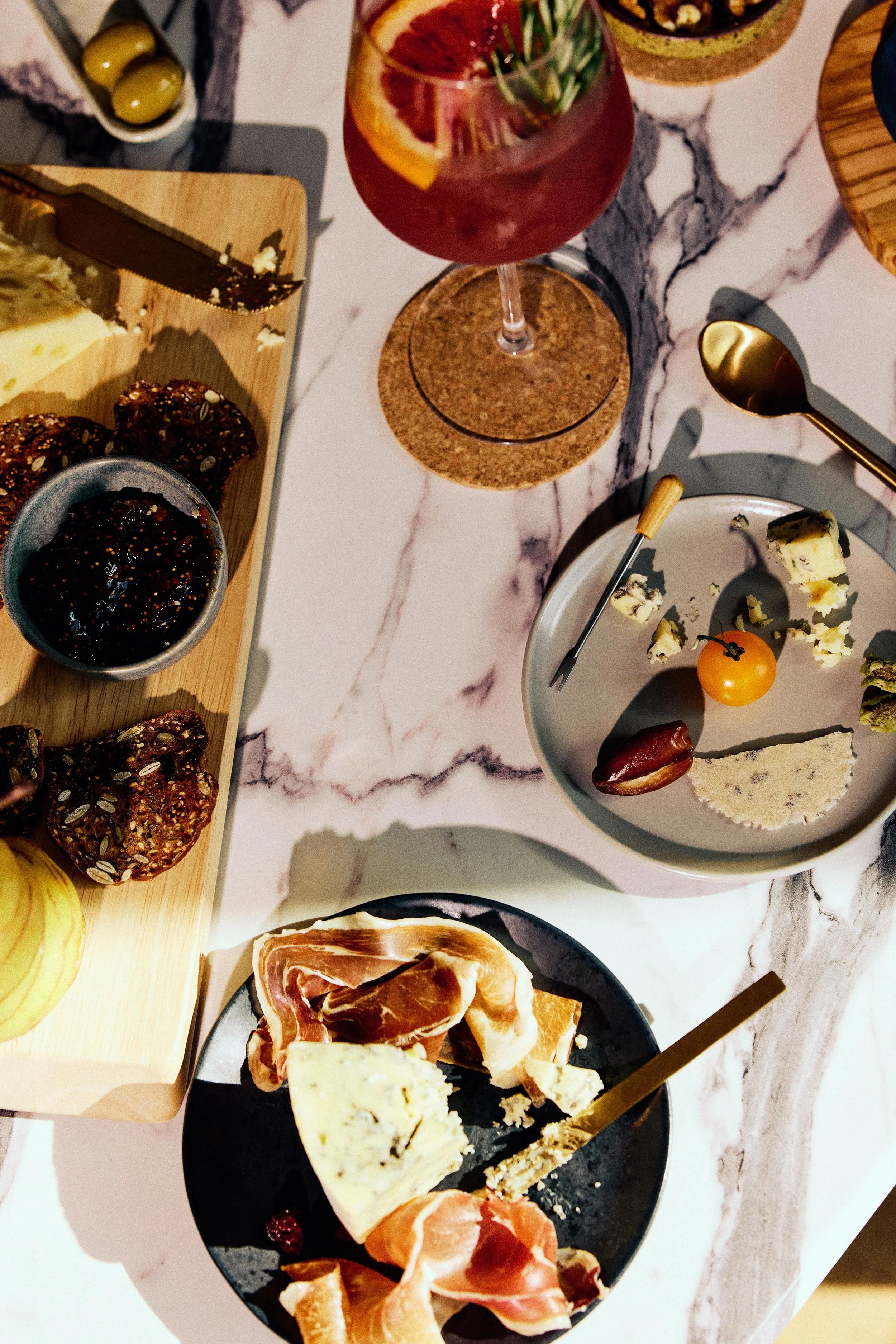 A charcuterie board with various cheeses, cured meats, a cherry tomato, and crackers, a glass of red drink with ice and fruit slices, and a small bowl of fig jam on a marble table.
