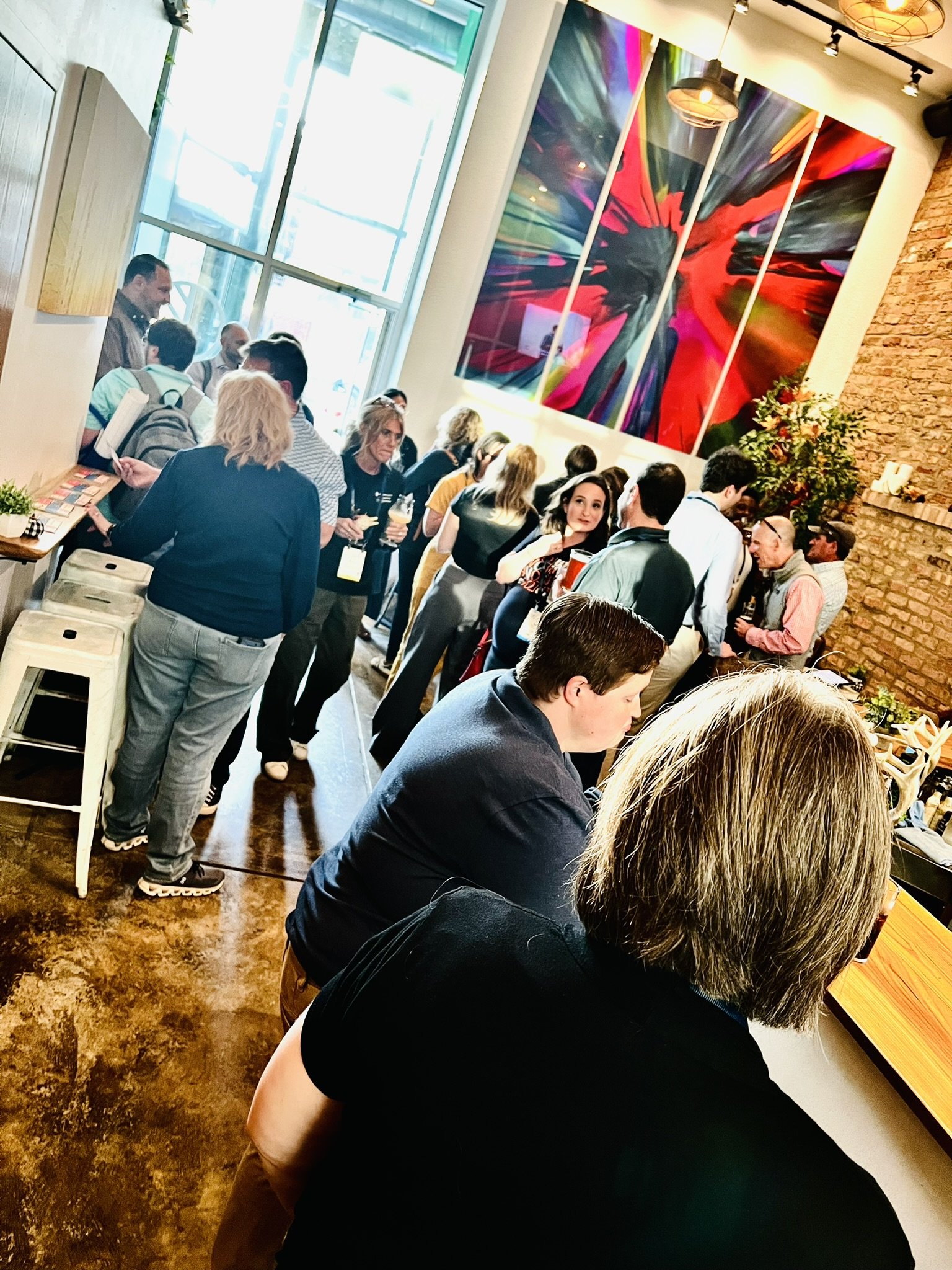 A crowded indoor event with people standing and socializing in a room featuring large colorful artwork on the wall, a brick wall, and a large window allowing natural light.