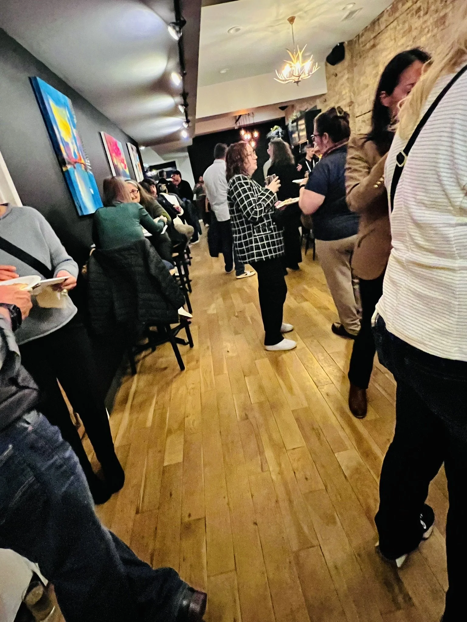 People standing in line inside a restaurant or bar, some holding plates or drinks, with colorful artwork on the wall and a chandelier hanging from the ceiling.