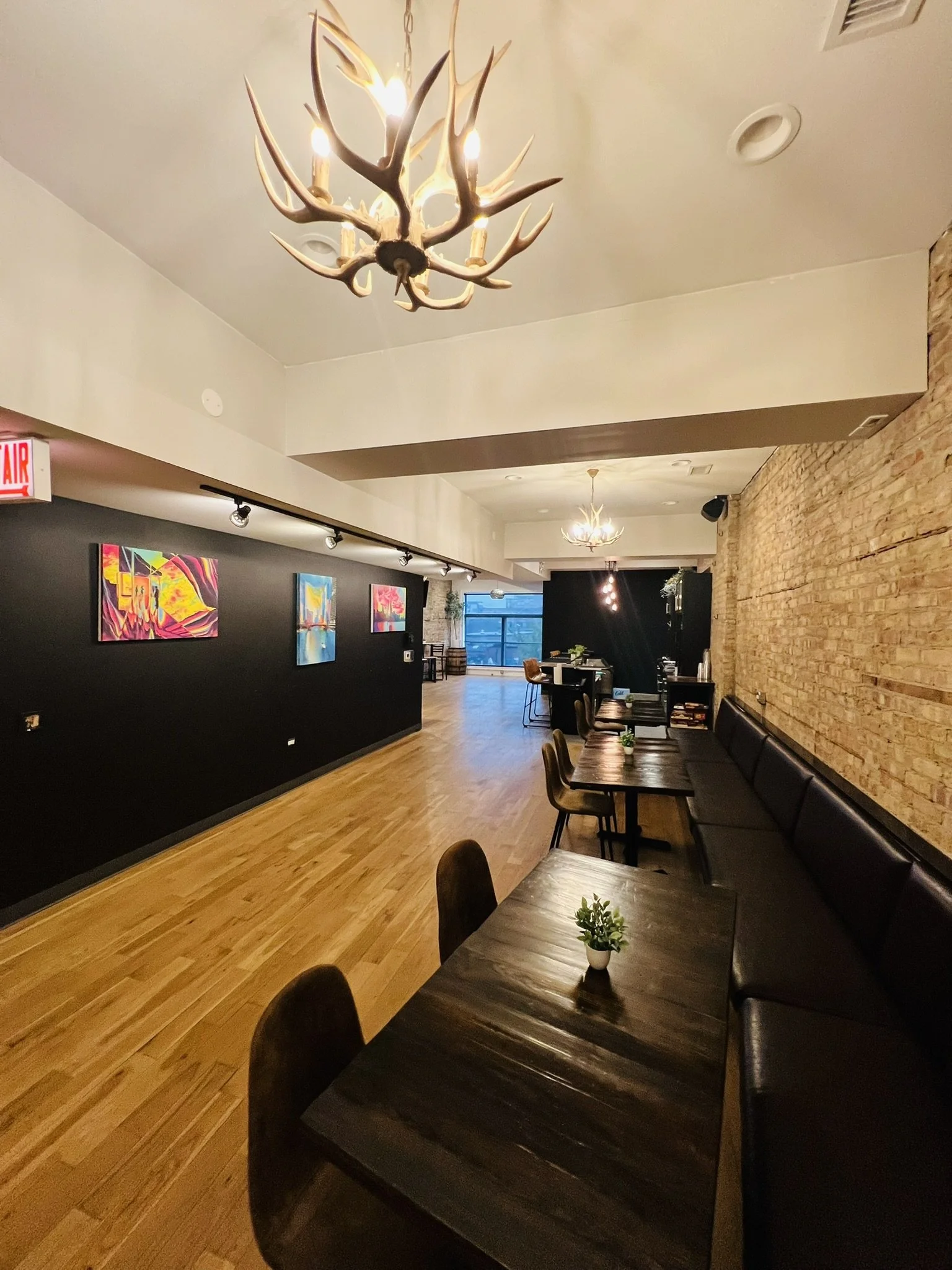 Interior view of a modern dining and lounge area with wood flooring, a brick wall, black benches and chairs, small potted plant on table, colorful artwork on black wall, antler chandelier, hanging pendant lights, and large windows at the far end.