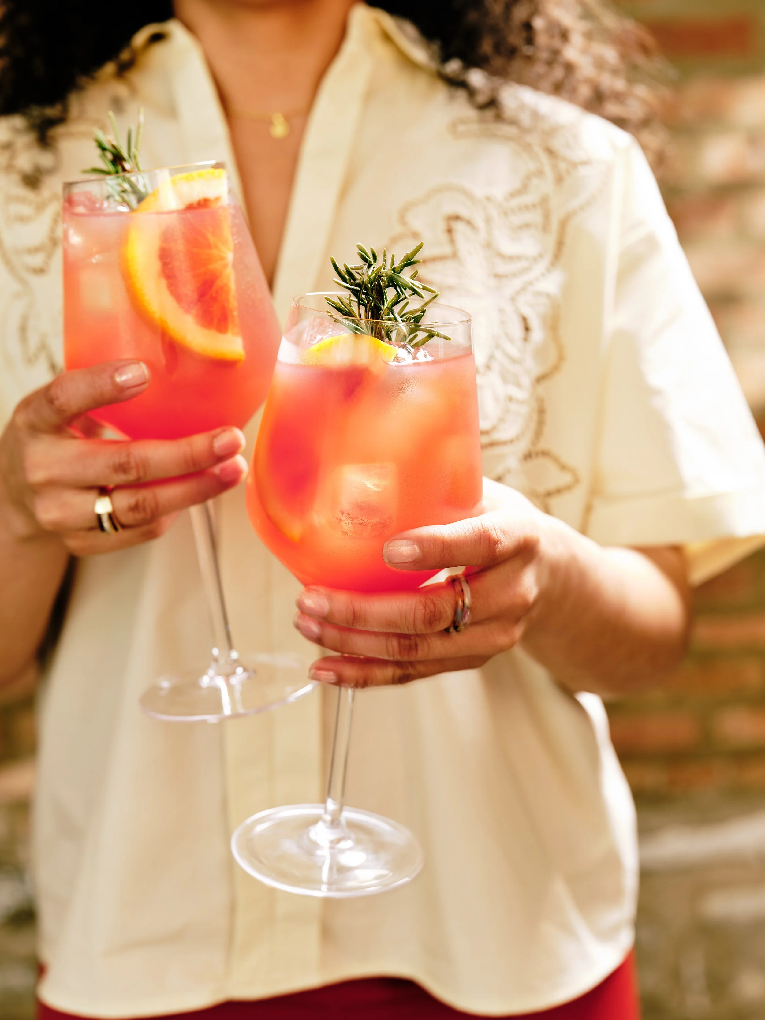 A person holding two pink cocktails garnished with orange slices and sprigs of rosemary, wearing a cream-colored embroidered blouse at Tethered Events Chicago