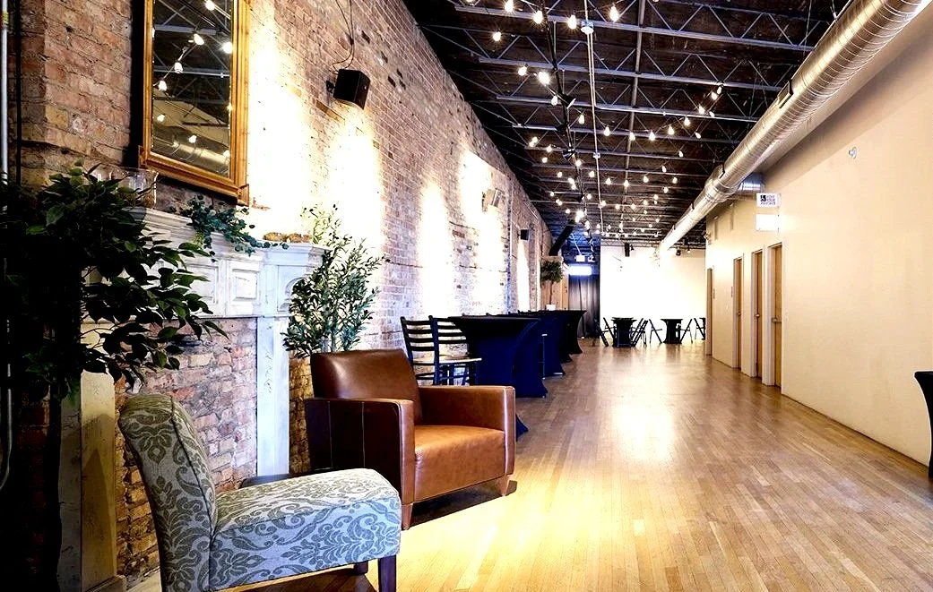 Spacious event space interior with wooden floor, exposed brick wall, string lights on ceiling, leather and patterned chairs, tall plants, and a long hallway.