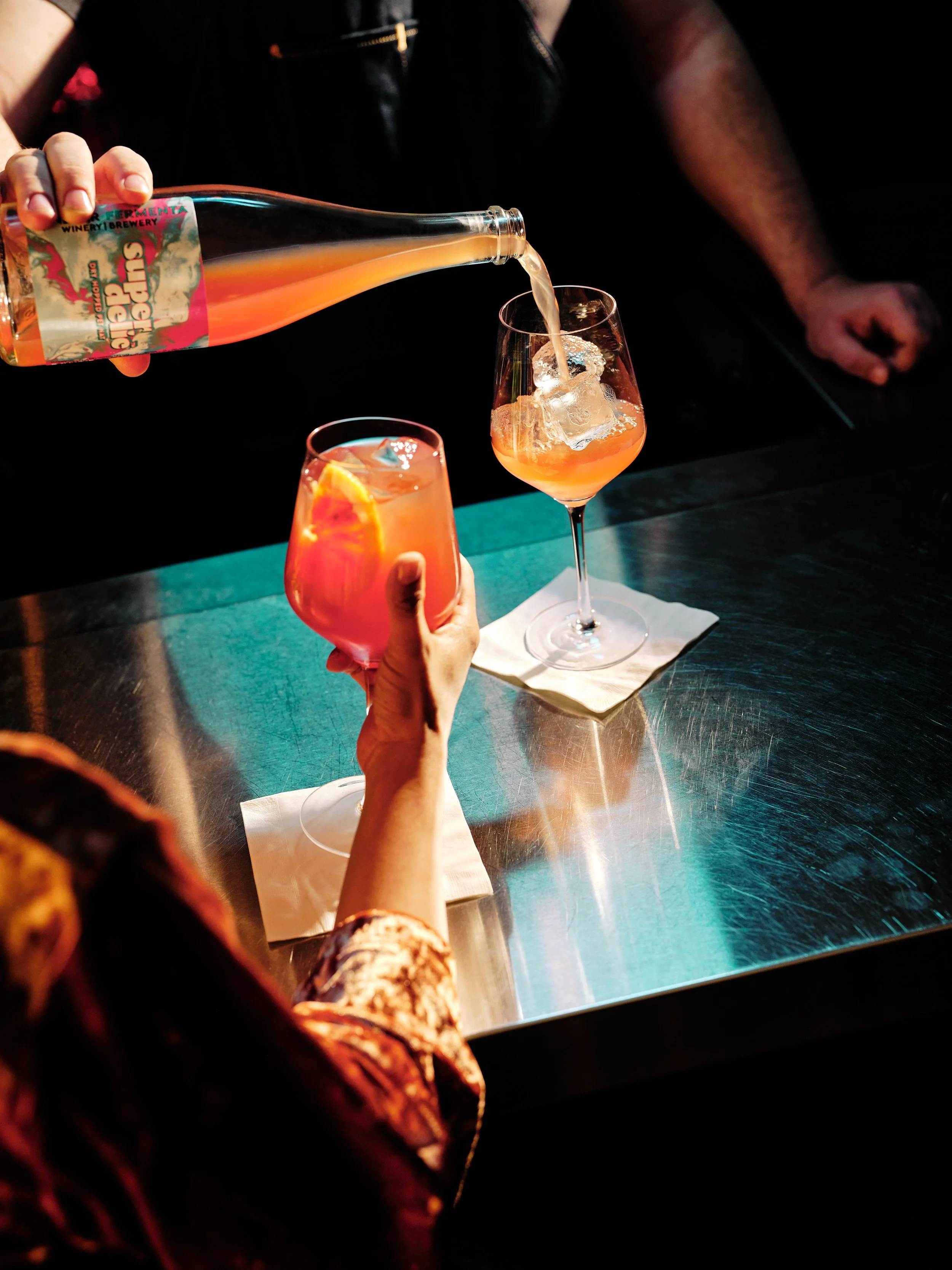 A person pouring a pinkish-orange cocktail into a glass with ice and a slice of orange, while another glass of similar drink with ice is on a napkin on a metallic table at Tethered Events