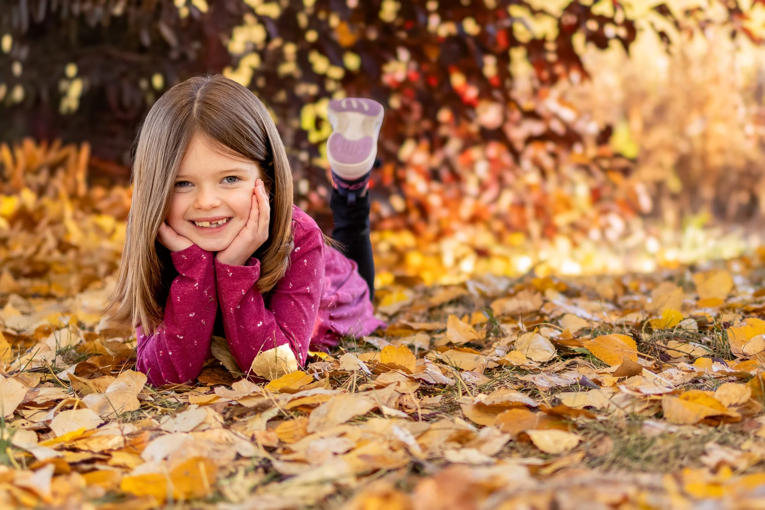 Little girl laying in golden fall leaves, smiling brightly during a family session in Airdrie.