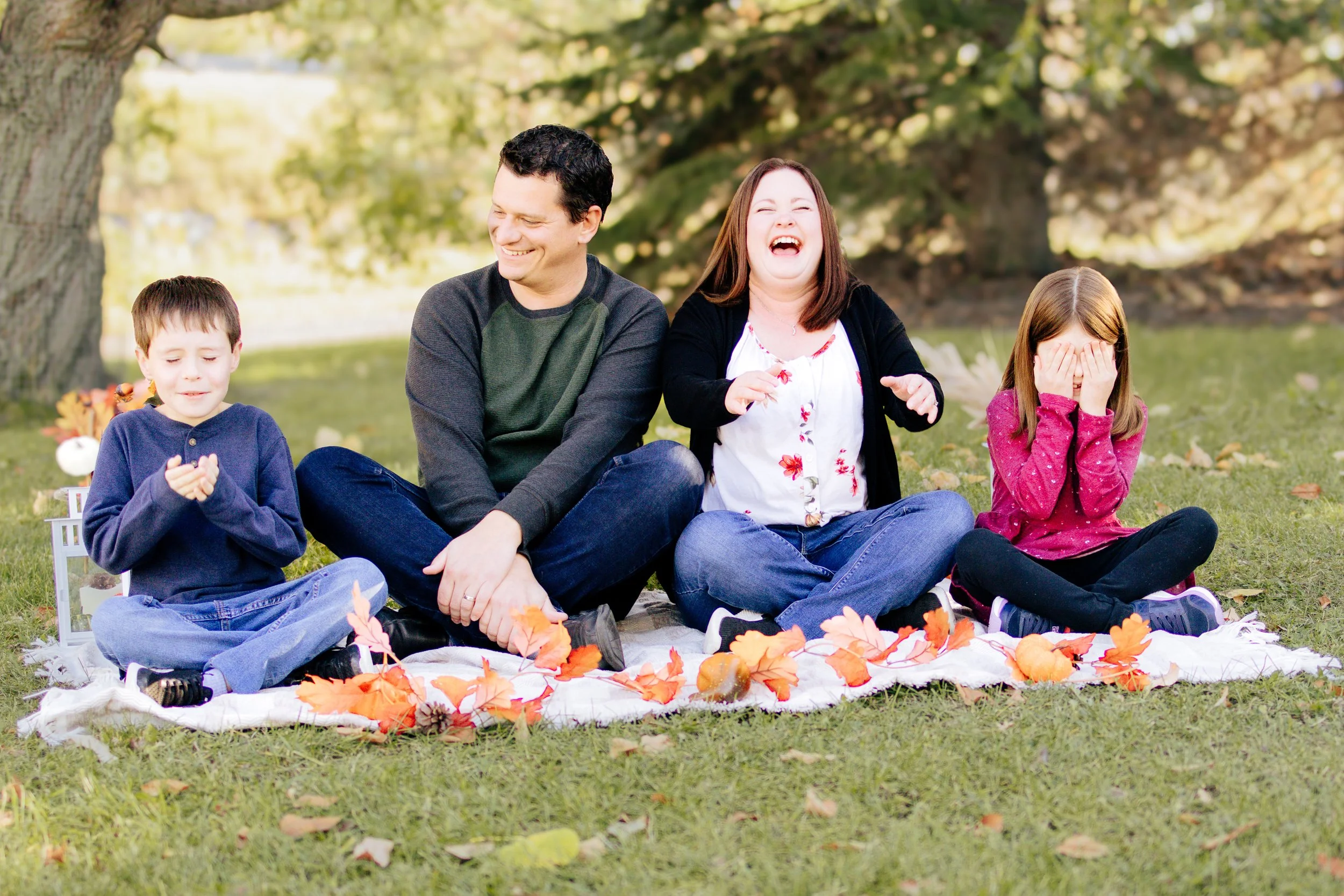 Family sitting together on a blanket in a park, surrounded by trees with changing leaves.
