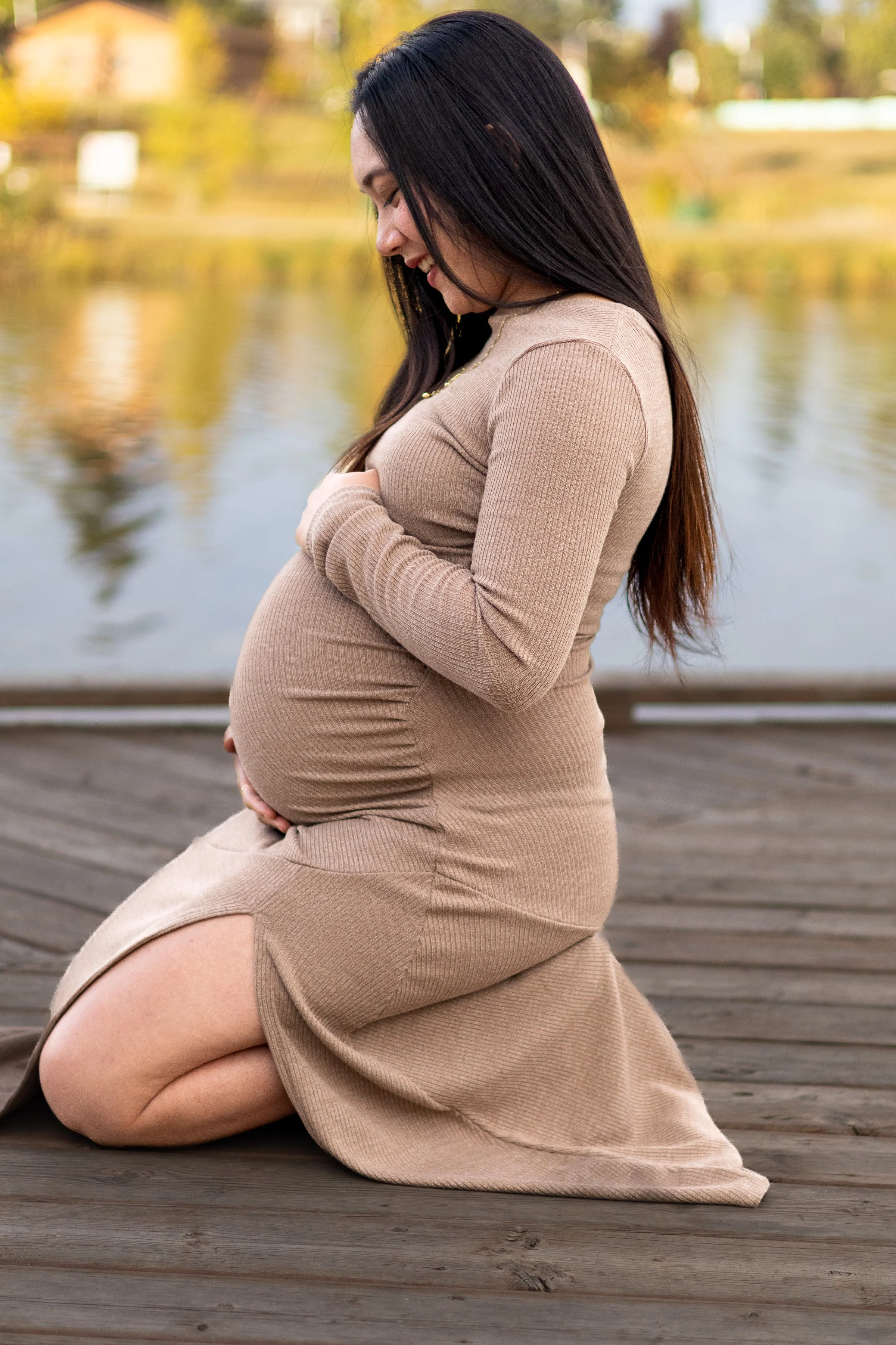 Pregnant woman kneeling on a wooden dock at Nose Creek Park in Airdrie, holding her belly with golden fall trees reflecting in the water.