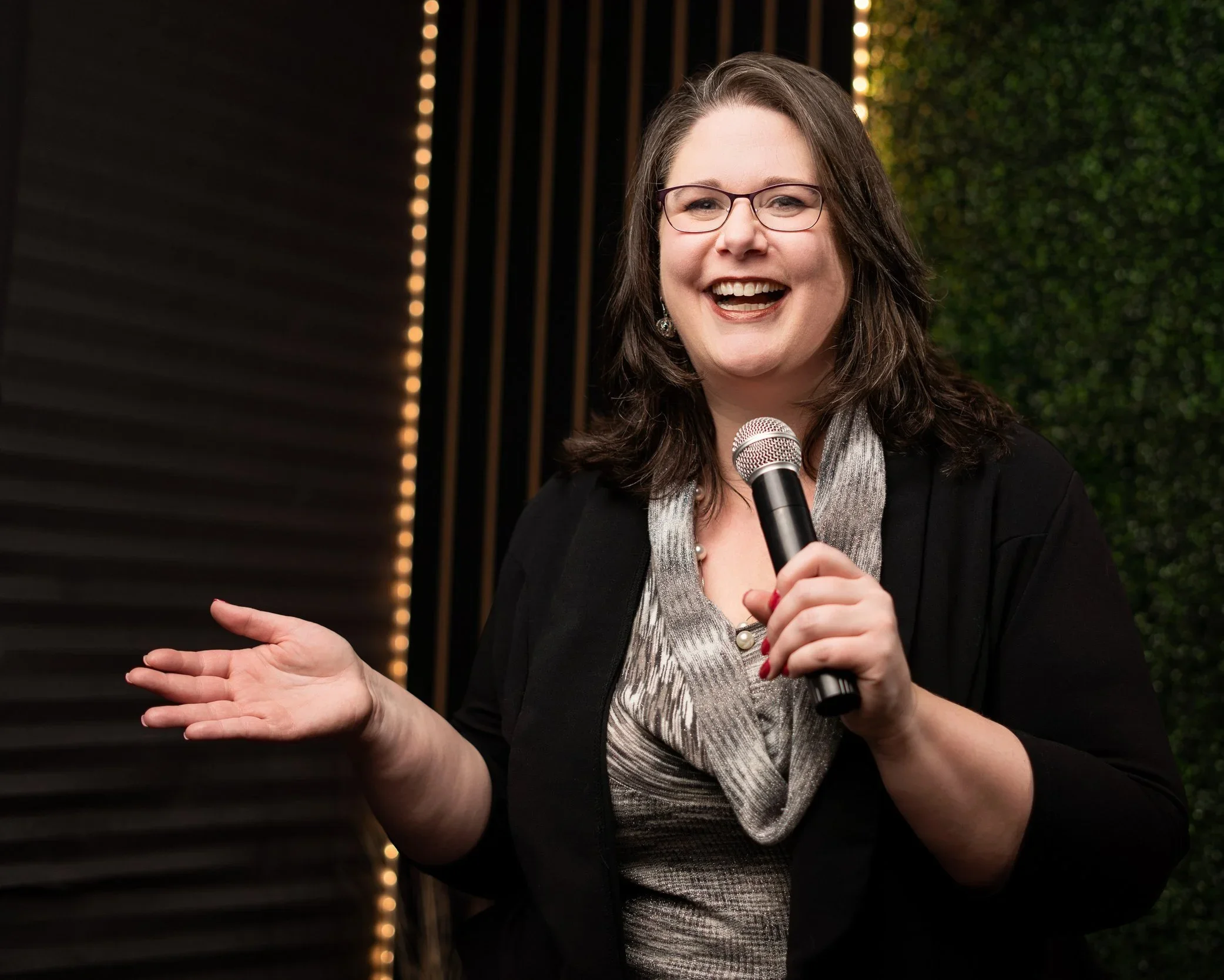 A woman with shoulder-length brown hair and purple lipstick standing outdoors against a brick wall, wearing a plaid jacket and a black T-shirt with purple and white graphic text.