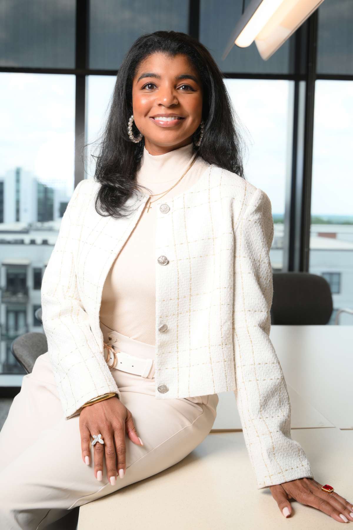 Close-up photo of a woman with straight, shoulder-length brown hair, wearing earrings, a white collared shirt, and a dark sweater, smiling softly indoors with warm lighting.