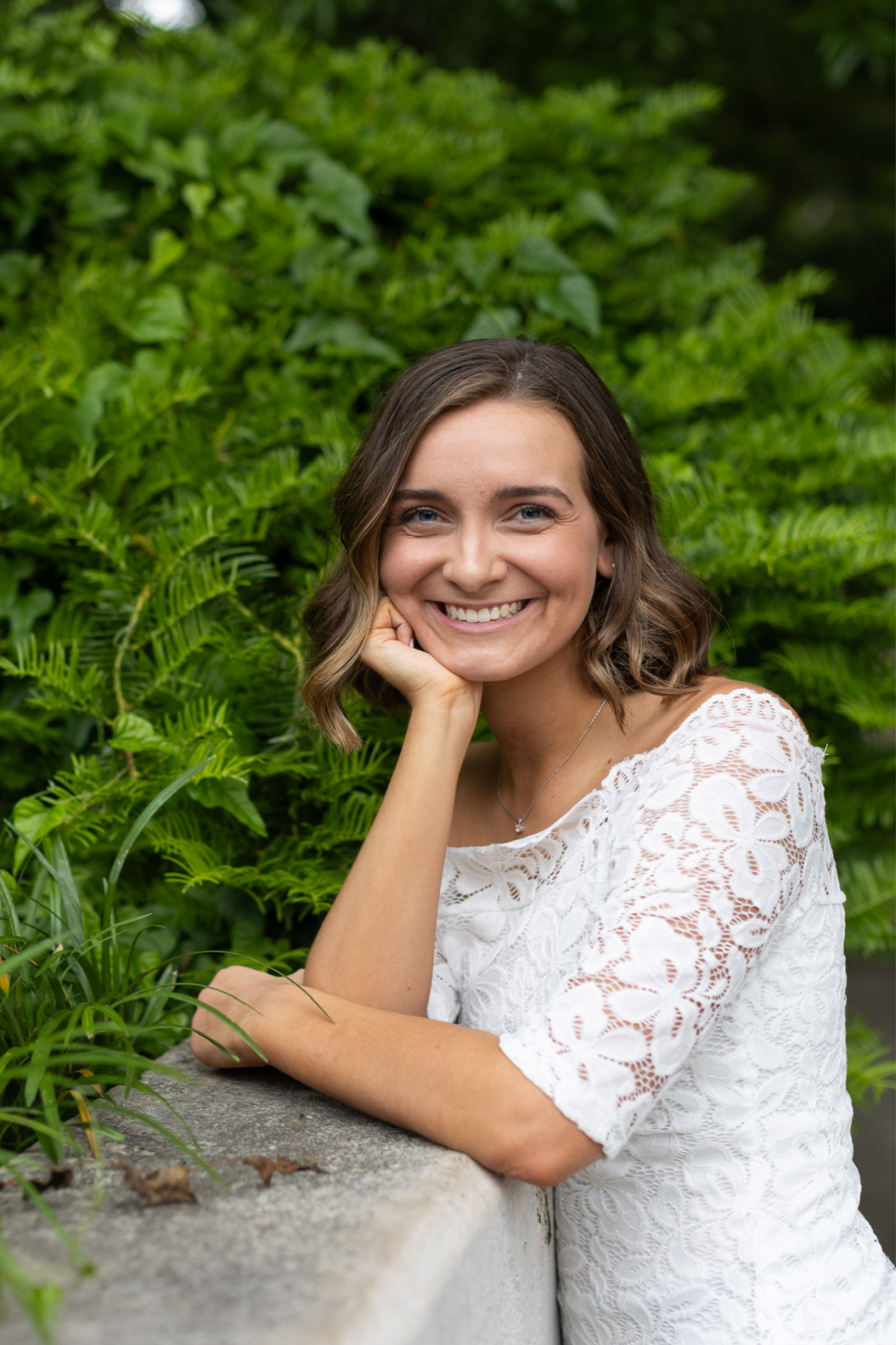 Black and white photo of a woman with dark hair, tattoos on her arm, wearing a t-shirt and a necklace, smiling slightly and resting her head on her hand.