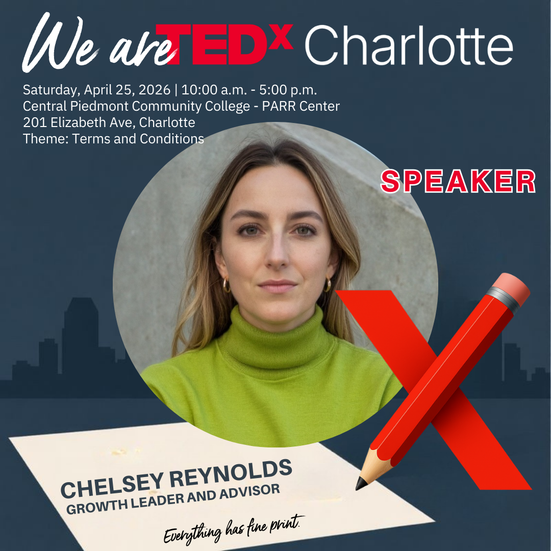 A woman with short curly hair and a bright smile, wearing a black blazer and a colorful patterned blouse, standing indoors with modern lighting and architecture in the background.