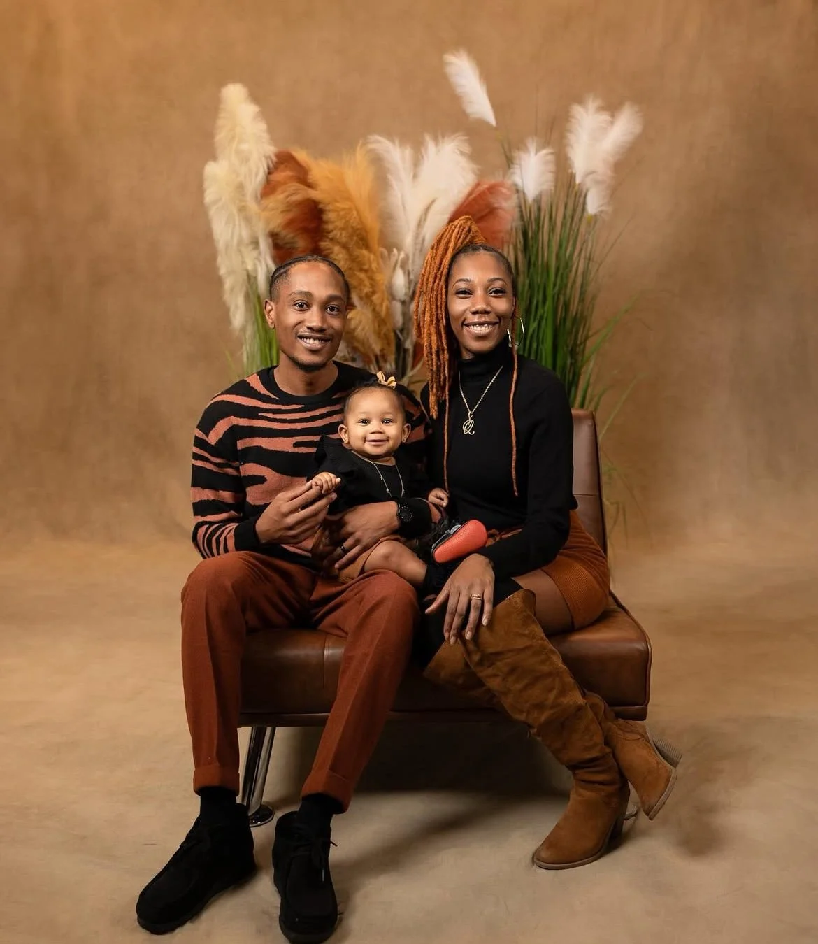 A happy family of three sitting on a brown leather bench, with a backdrop of tall pampas grass and dried flowers.