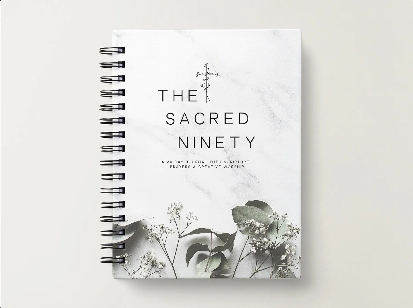 A spiral-bound journal titled "The Sacred Ninety" with a subtitle "A 30-day journal with scripture, prayers & creative worship". The cover features a minimalist design with a small illustration of a plant and a few sprigs of white flowers and green leaves at the bottom.
