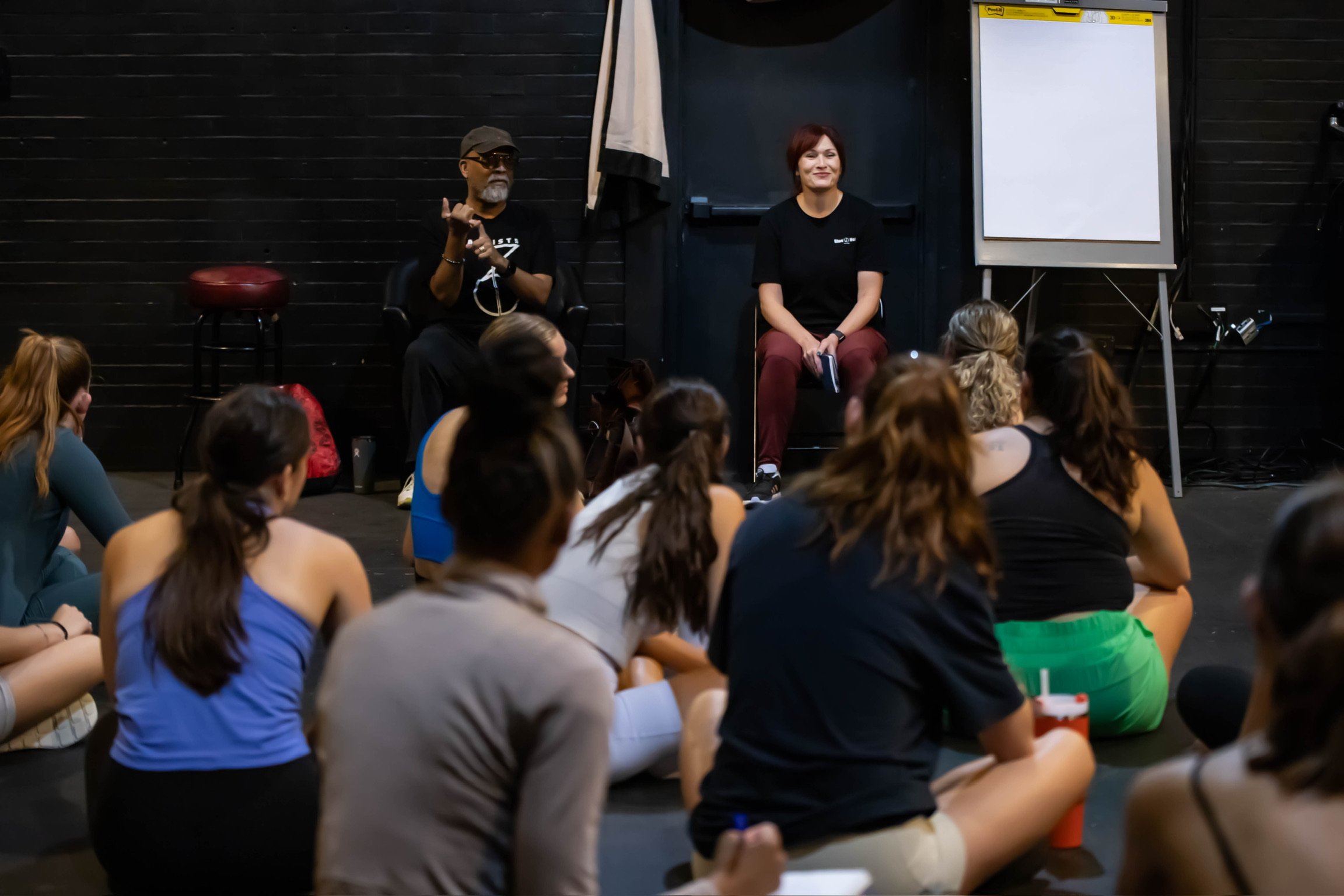 A group of women sitting on the floor listening to an instructor and a speaker at a workshop or class in a dimly lit room with black walls, with a whiteboard on a stand nearby.