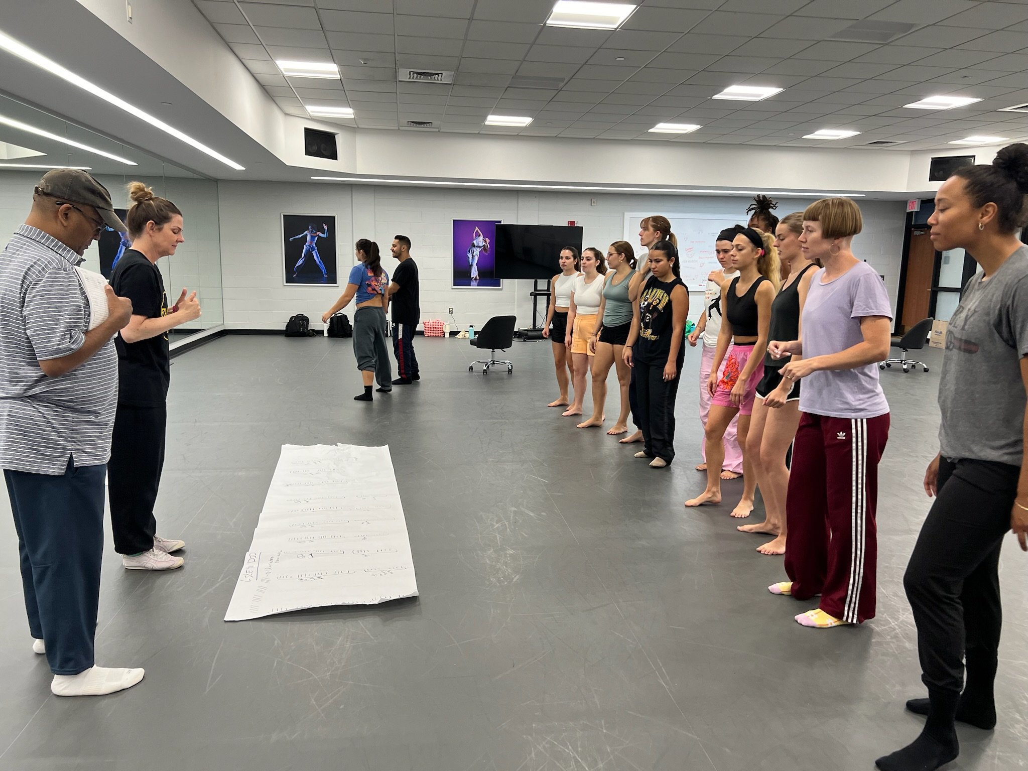 A group of dancers standing in a line in a dance studio, listening to instructors while wearing dance attire.