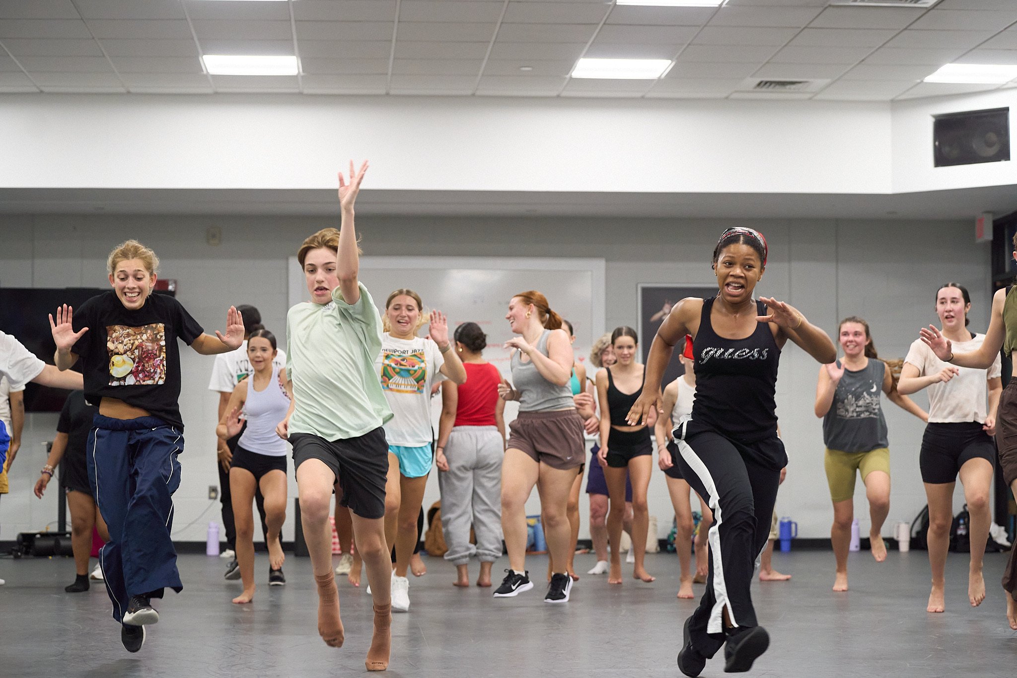 Group of people, mostly women, practicing dance in a spacious, indoor studio. They are mid-movement, smiling, and wearing athletic or comfortable clothing.