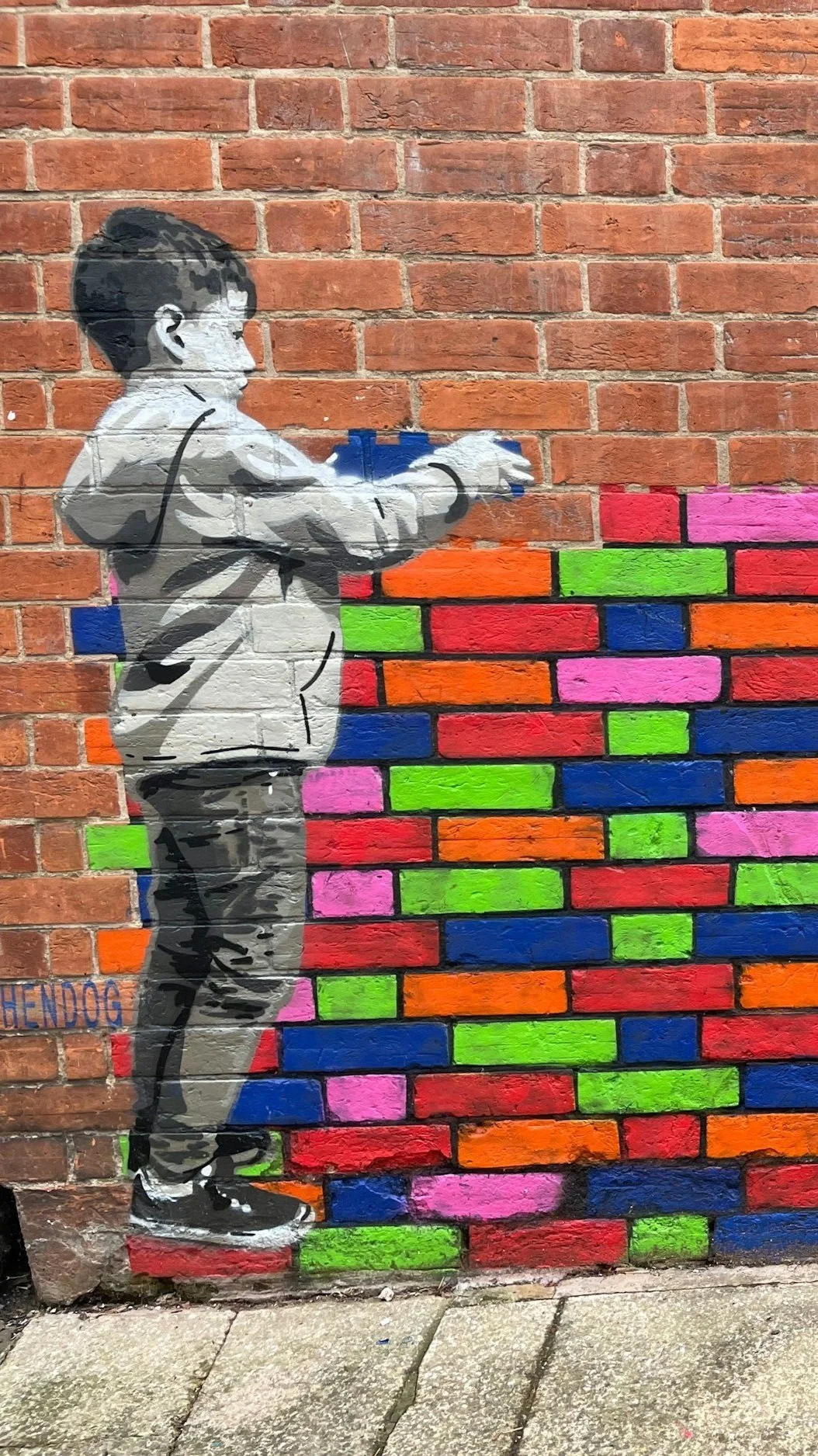 Street art of a young boy in a hoodie and pants spray painting colorful bricks on a brick wall.