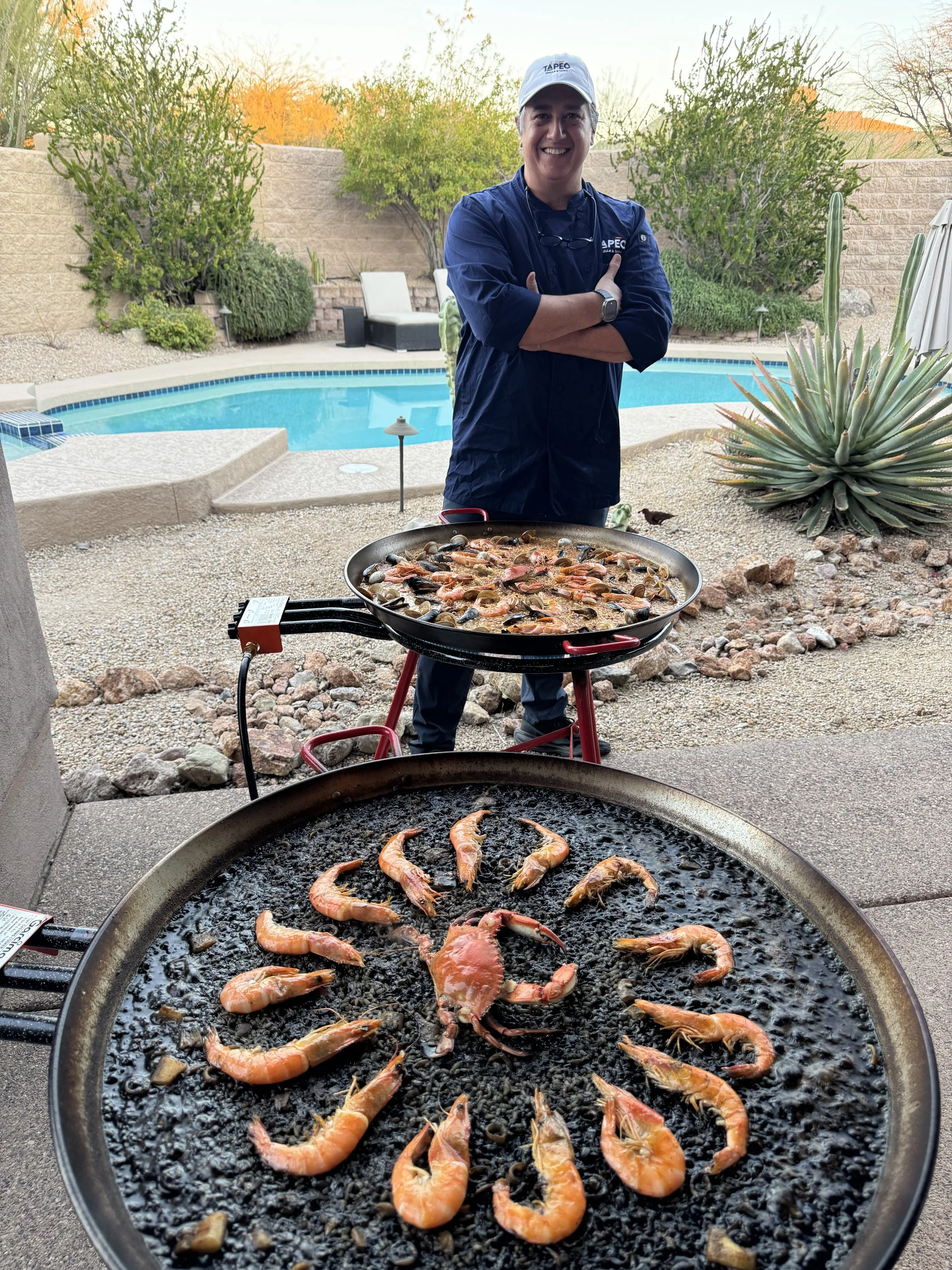 A smiling chef standing outside near a swimming pool with a backyard garden, with two grills in foreground cooking shrimp and crab.