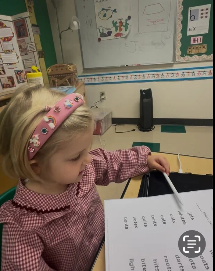 A young girl with blonde hair, wearing a pink headband decorated with unicorns and rainbows, sitting at a table in a classroom, working with a pointer on a worksheet with printed words.