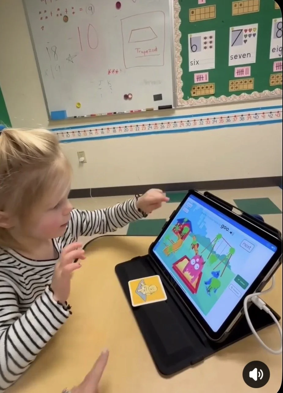 A young girl with blonde hair, wearing a black and white striped shirt, seated at a desk in a classroom. She is interacting with a tablet that displays an educational game involving colorful characters and a castle. A playing card with a yellow duck image is on the desk next to the tablet. In the background, there is a whiteboard with math doodles, a number line, and a bulletin board with number and shape posters.