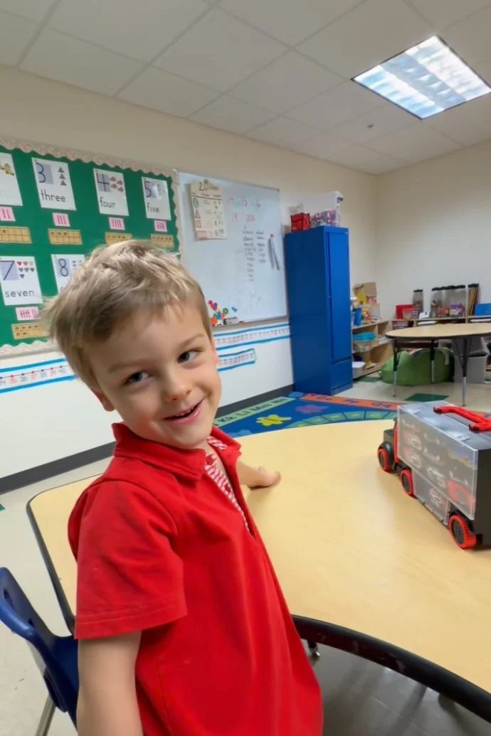 A young boy with light brown hair smiling, wearing a red shirt, standing near a table in a classroom.