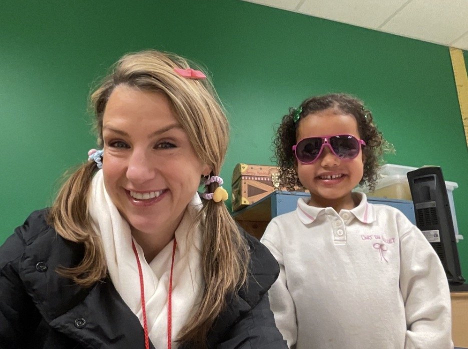 A young woman and a girl smiling for a selfie in a classroom. The woman has blonde hair in pigtails with colorful hair ties and a pink clip, and the girl has curly hair with pink sunglasses and a white shirt.