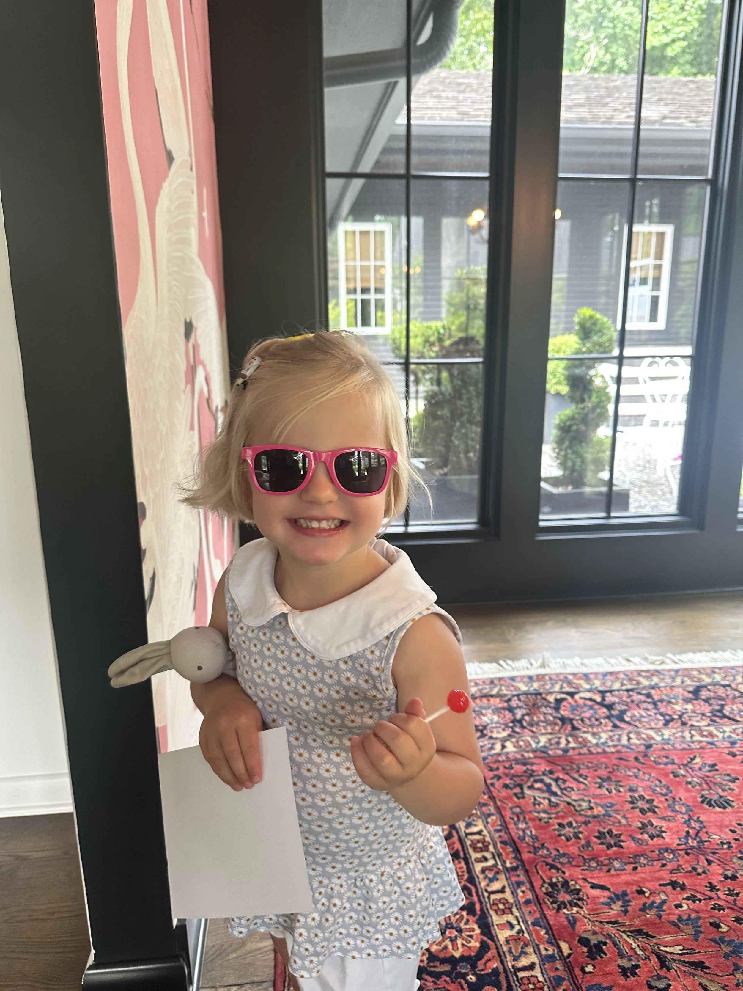 A young girl with blonde hair wearing pink sunglasses, a daisy print dress with a white collar, holding a red lollipop in one hand and a stuffed bunny toy in the other, standing indoors near a large window with black frames and a patterned rug on the floor.