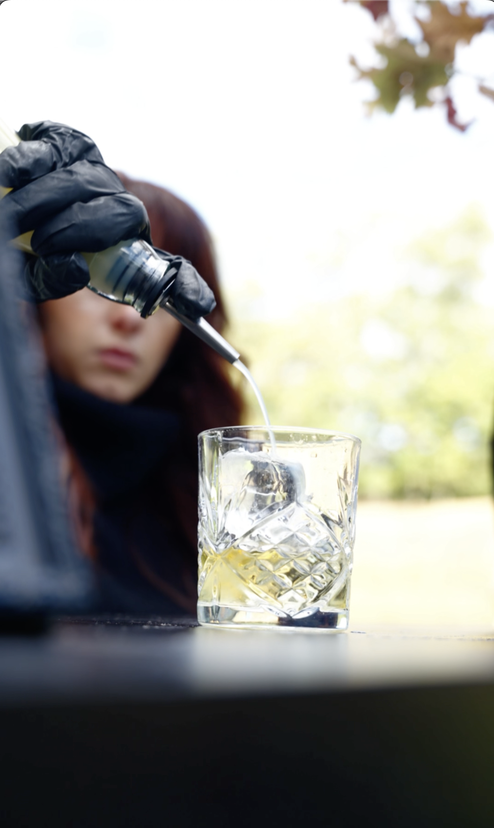 A close-up of a cocktail with ice cubes, being poured with a dark spirit, garnished with a lemon twist on the glass rim, on a wooden surface.