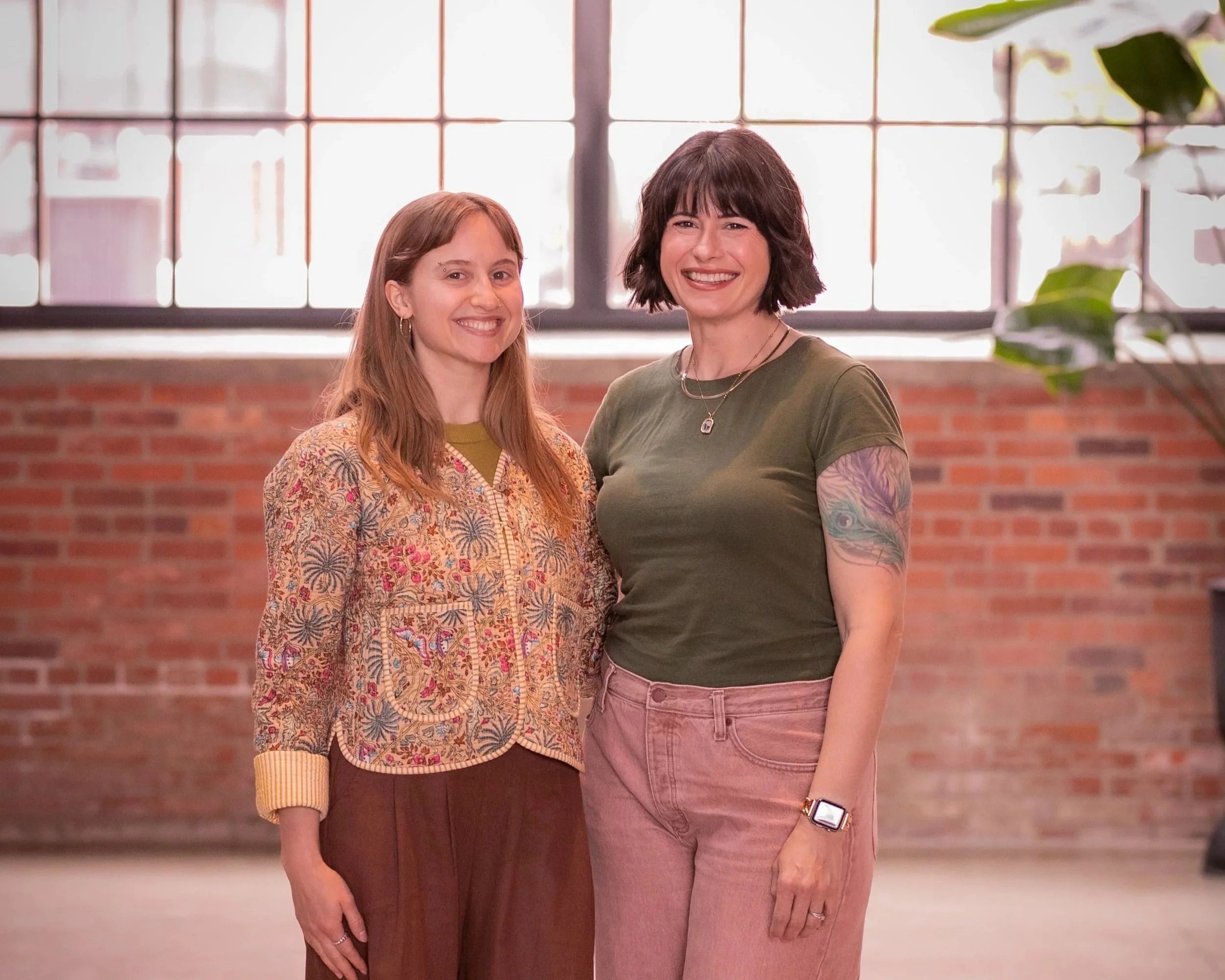 Two women standing in front of a large window, smiling at the camera. One woman has long red hair and wears a floral patterned top, while the other woman has short dark hair, a tattooed arm, and is dressed in a green t-shirt and pink pants.
