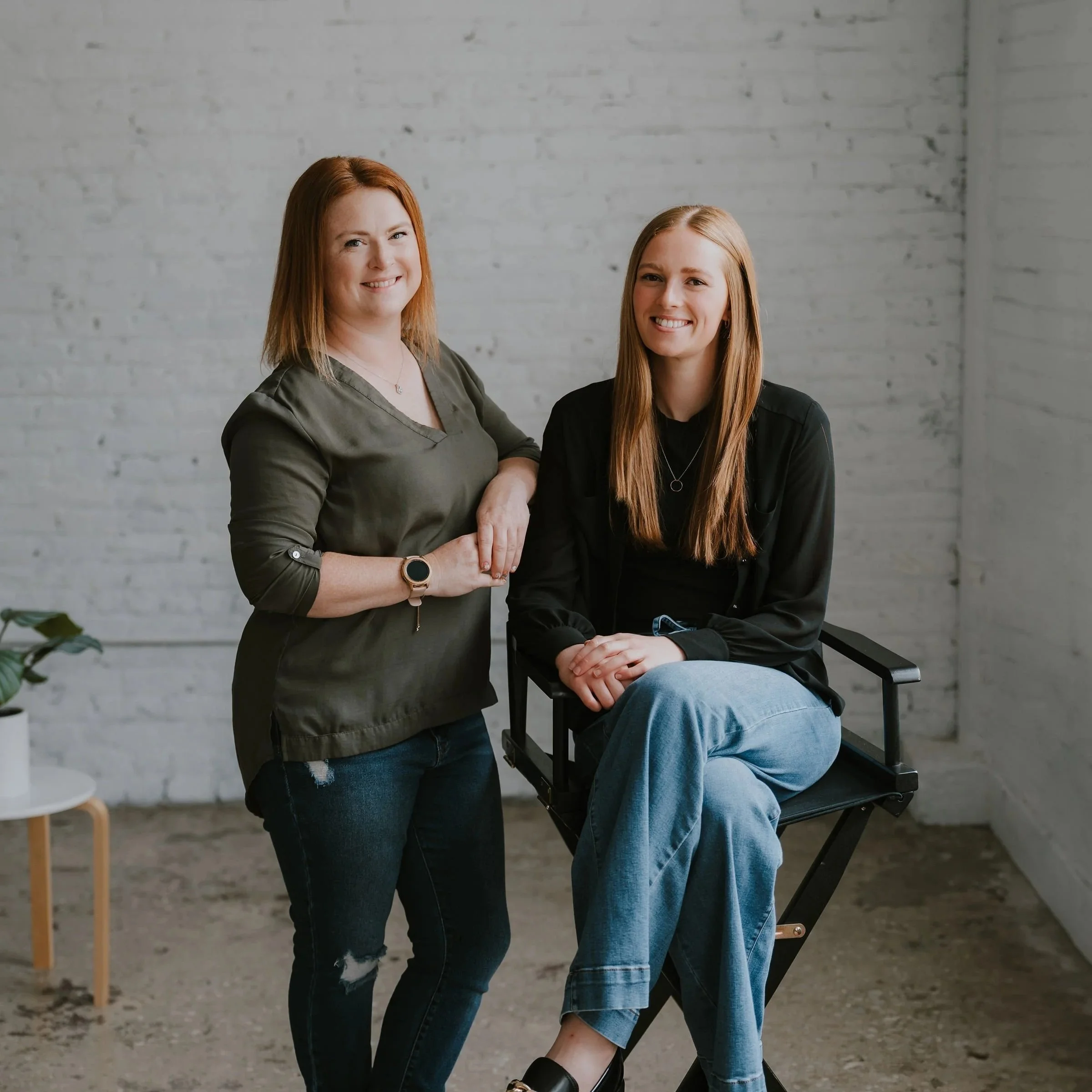 Two women with long hair smiling, one with red hair and the other with light brown hair, in casual clothing, posing together in front of a white brick wall. One is standing, and the other is seated on a black chair.