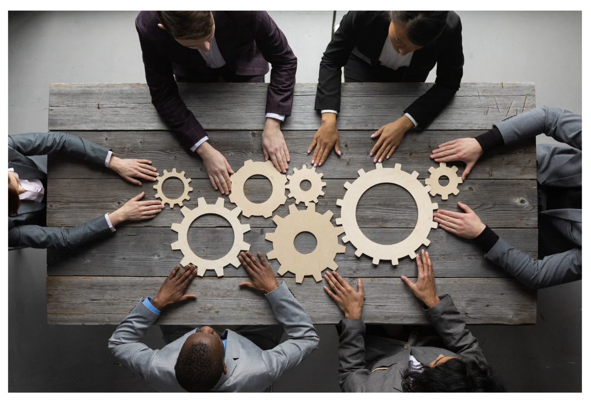 Six businesspeople gathered around a wooden table, working with wooden gear cutouts to symbolize teamwork or collaboration.