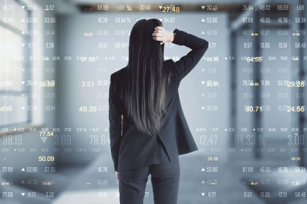 Businesswoman standing in front of a digital display of fluctuating stock market numbers and percentages, facing away with one hand on her head.