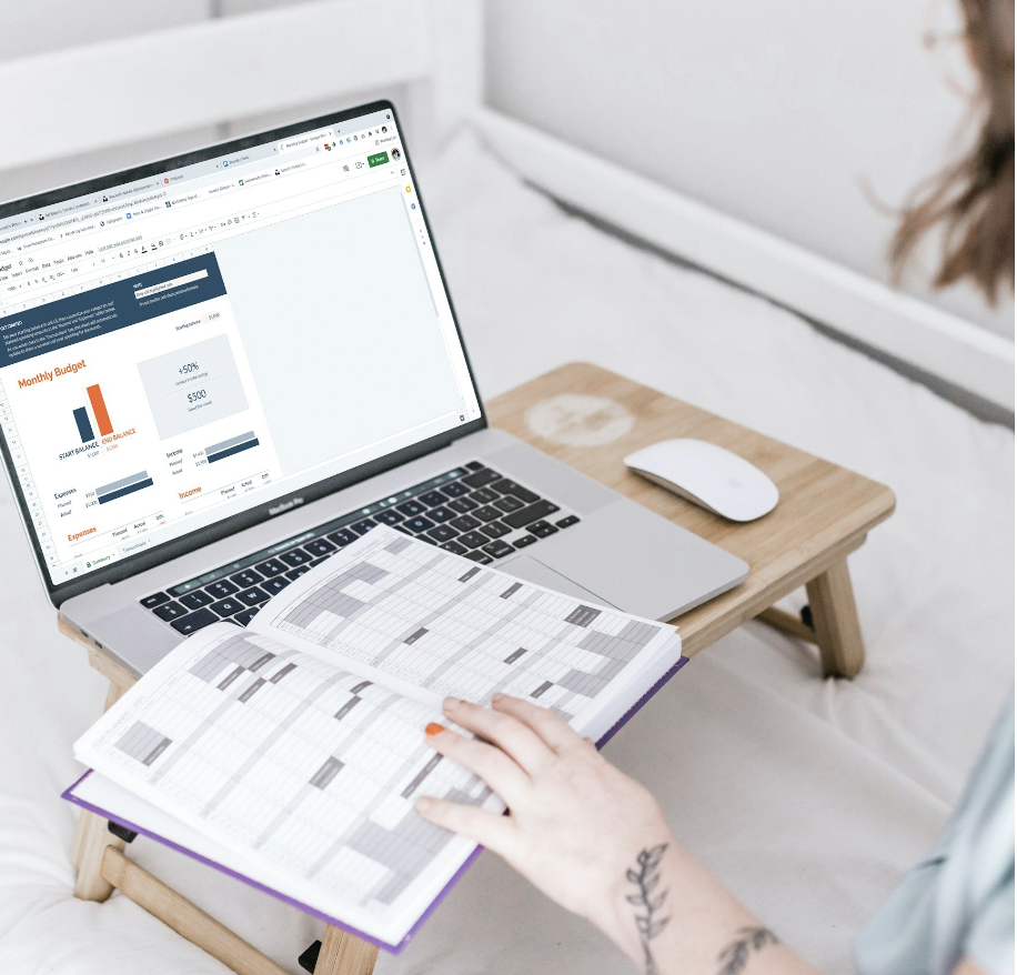 A person is working with a laptop, printed schedules, and a pen on a small wooden table. The laptop screen displays a financial graph and a budget spreadsheet.
