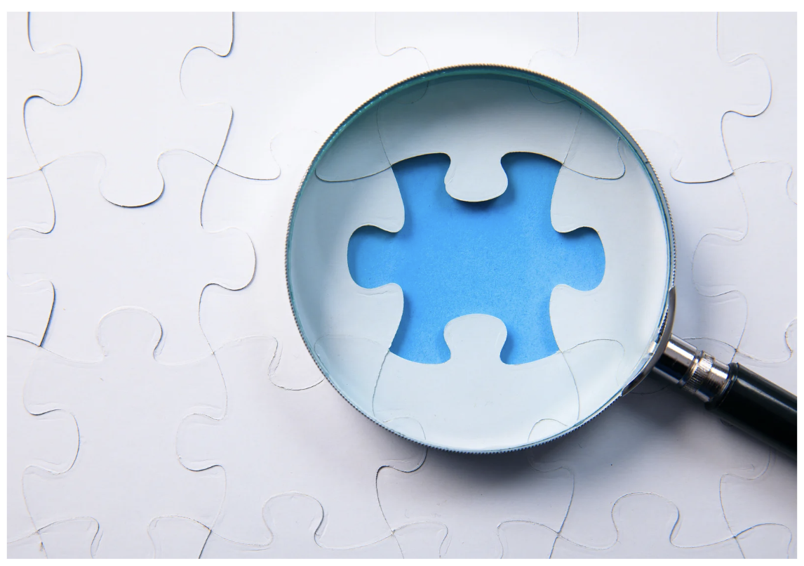 A partially assembled white jigsaw puzzle with a hole in the center, a magnifying glass focusing on the hole, revealing a blue background inside.