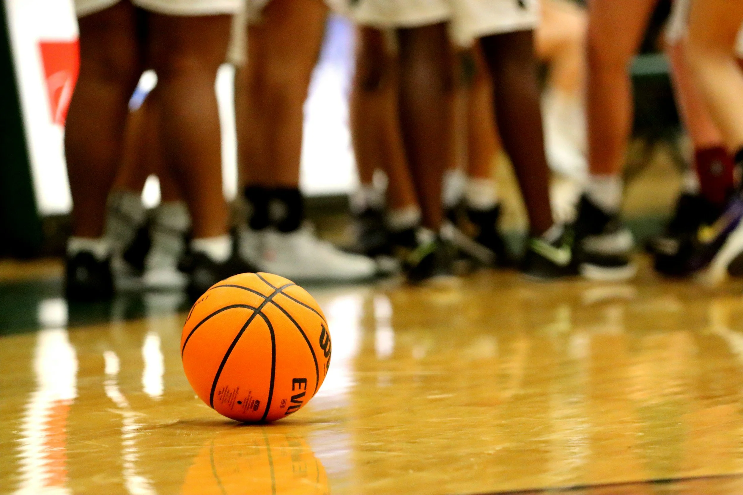 A yellow basketball resting on a shiny gymnasium floor with a group of kids and their legs in shorts and sneakers in the background.