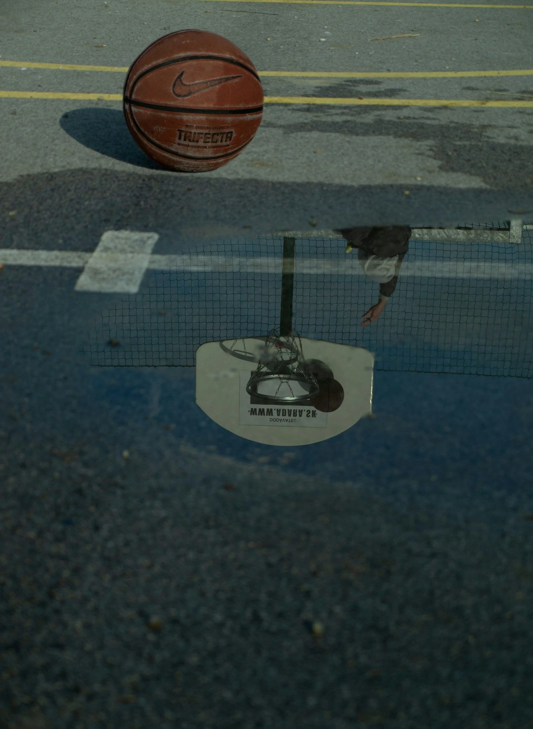 A basketball on a concrete court with yellow lines, reflected upside down in a puddle that shows a person reaching toward a basketball hoop.