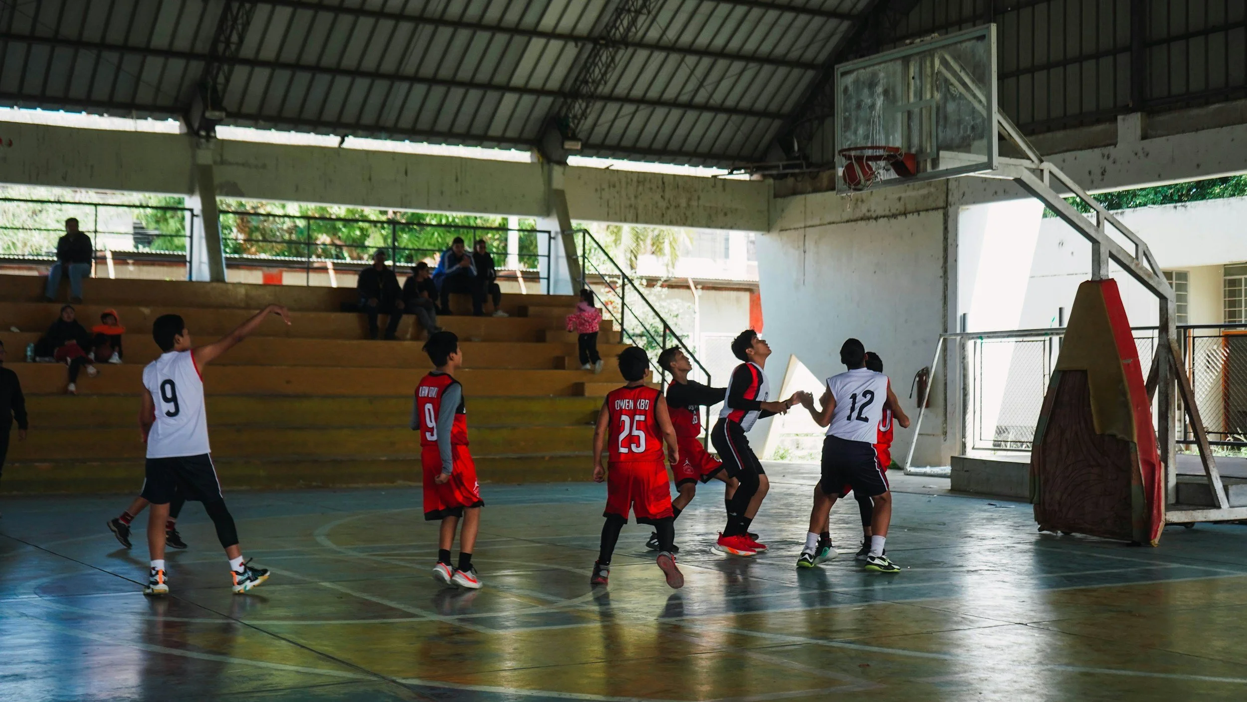 Kids playing basketball on an indoor court with spectators watching from the bleachers.