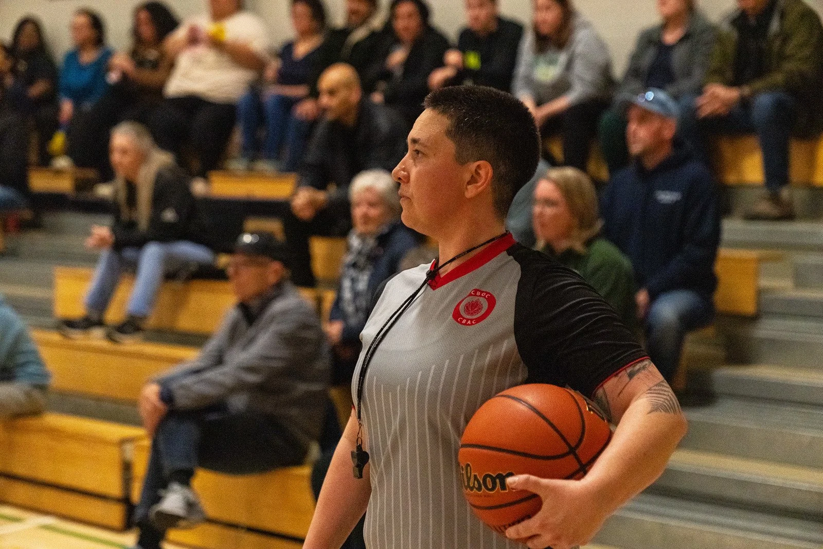 A female basketball player holding a Wilson basketball during a game or practice session, with spectators sitting on bleachers in the background.