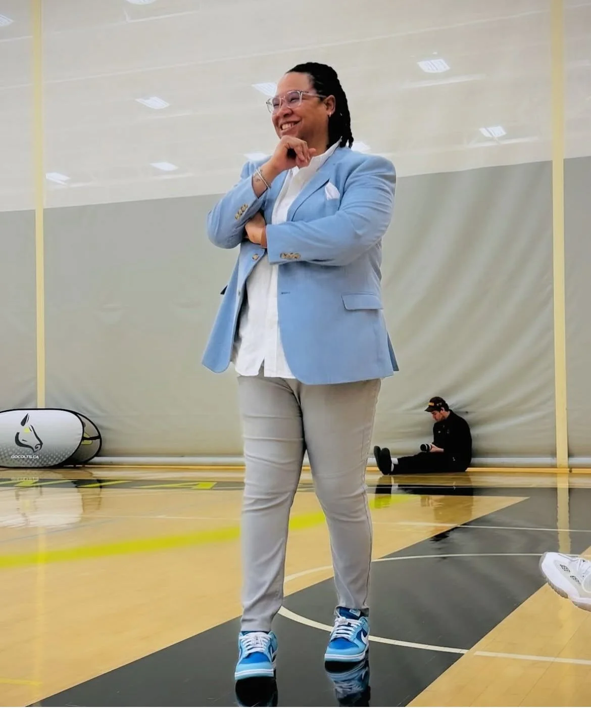A person in a light blue blazer, white shirt, and beige pants standing on a sports court, smiling with crossed arms, wearing blue sneakers, with a person sitting in the background.
