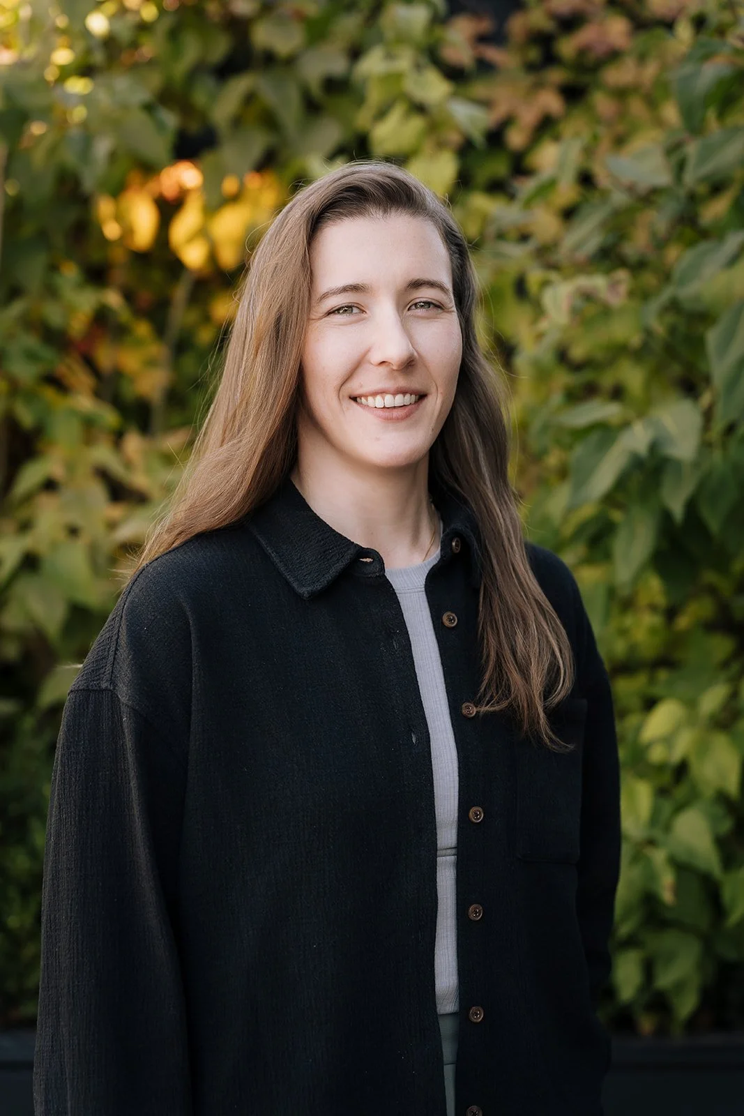 A young woman with long brown hair smiling outdoors, standing in front of green foliage.